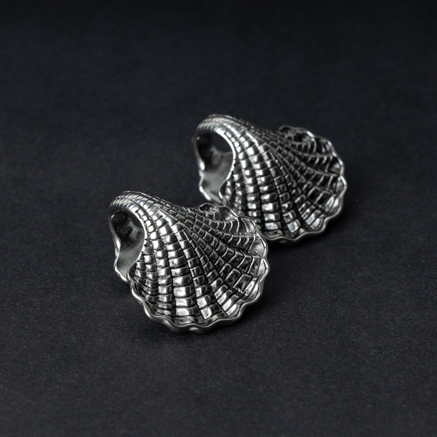 Two seashell-shaped ear weights in polished silver, with detailed textures and a bold design, displayed on a dark background.