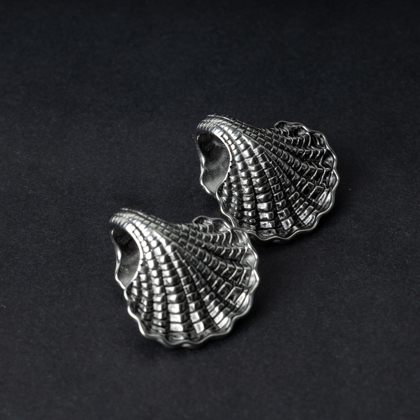 Two seashell-shaped ear weights in polished silver, with detailed textures and a bold design, displayed on a dark background.
