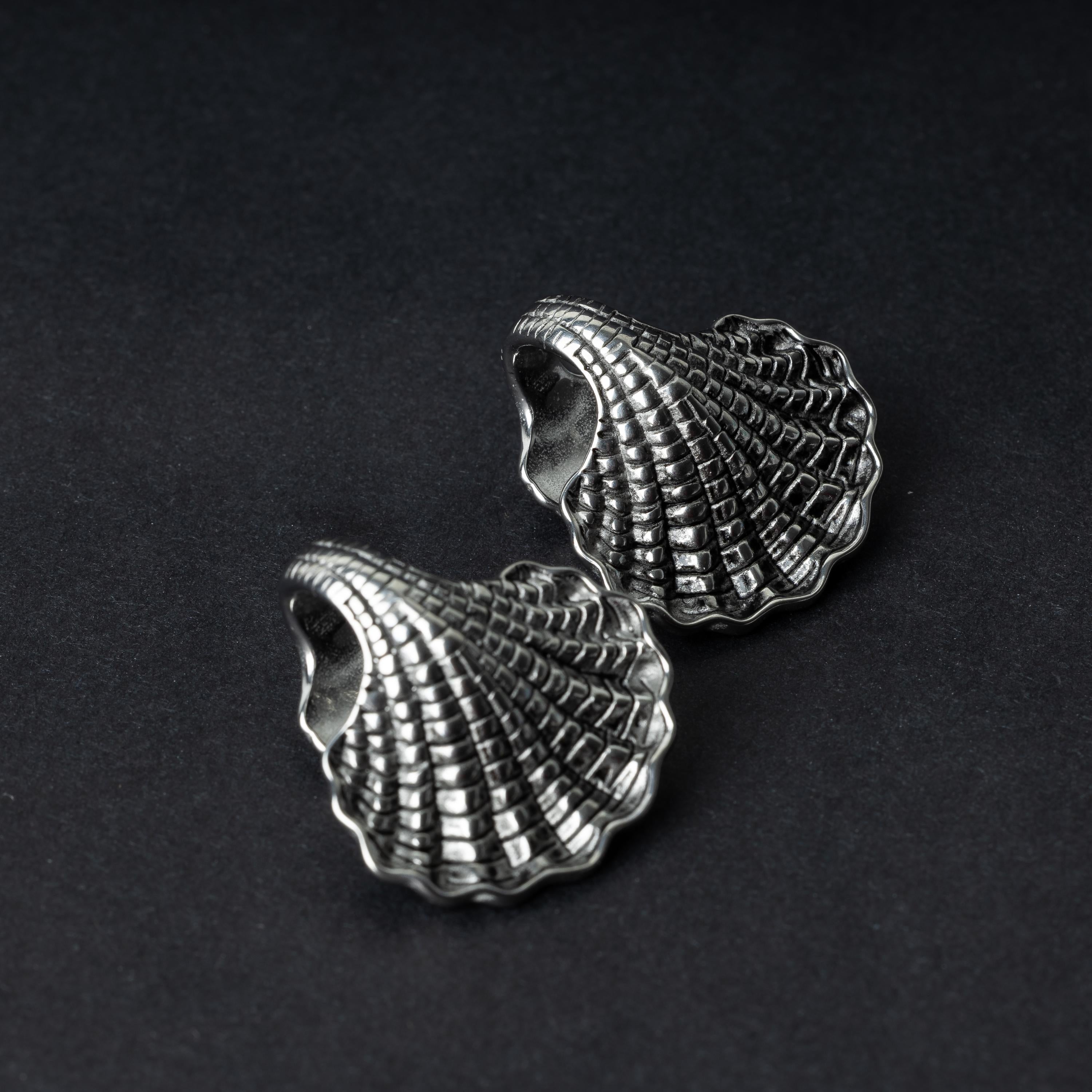 Two seashell-shaped ear weights in polished silver, with detailed textures and a bold design, displayed on a dark background.