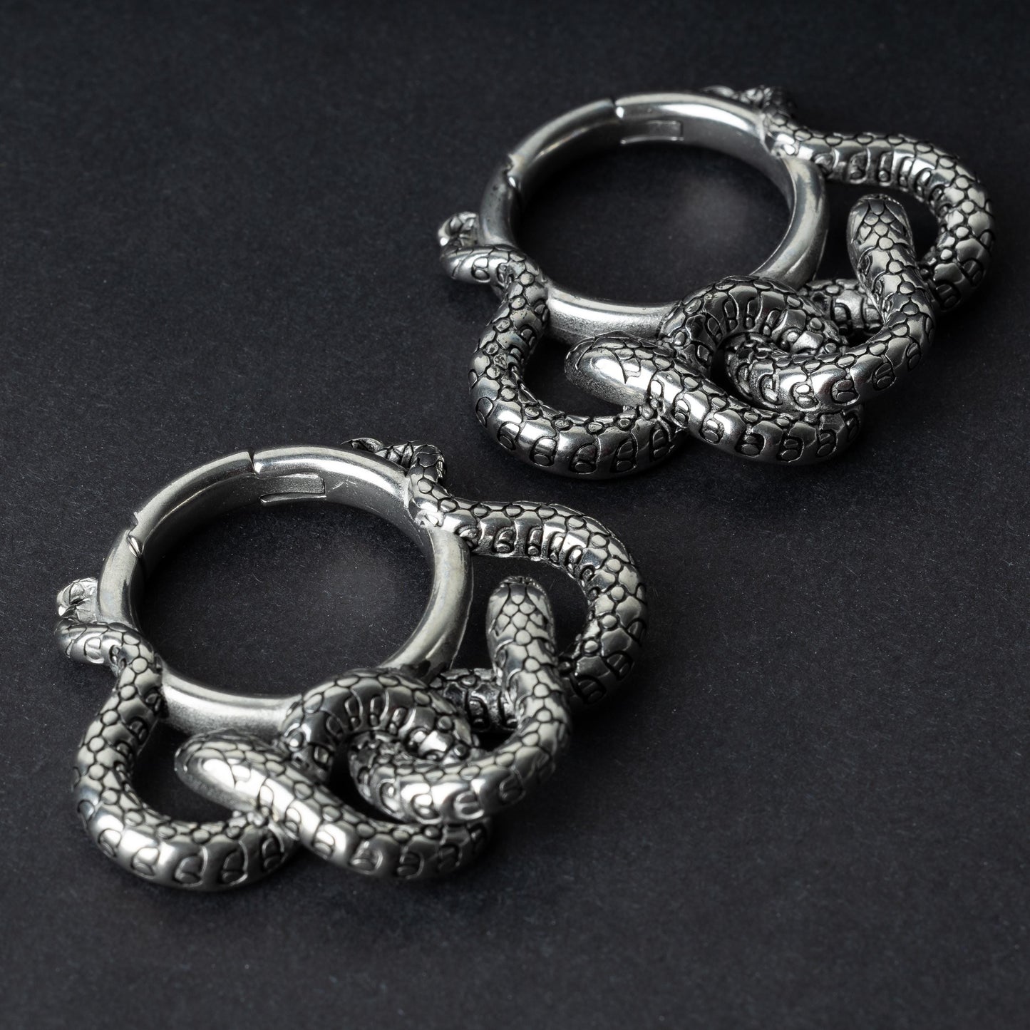 Two snake ear weights in polished silver, featuring detailed scales and a seamless hinge closure, displayed on a dark background.
