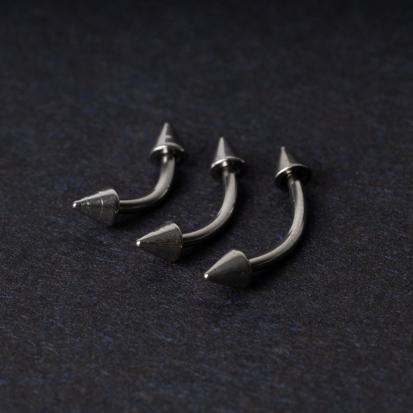 Three stainless steel curved barbells with cone-shaped ends, displayed on a dark background.