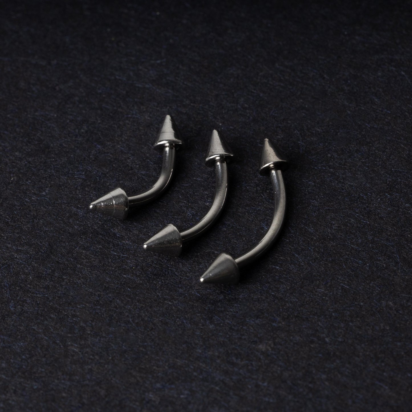 Three stainless steel curved barbells with cone-shaped ends, displayed on a dark background.