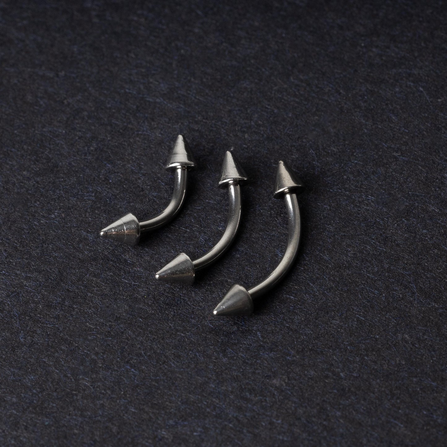 Three stainless steel curved barbells with cone-shaped ends, displayed on a dark background.