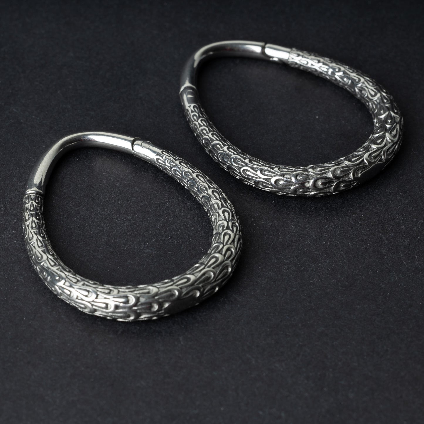 A pair of silver teardrop ear weights featuring a detailed dragon-scale texture and a seamless hinge closure, displayed on a dark background.
