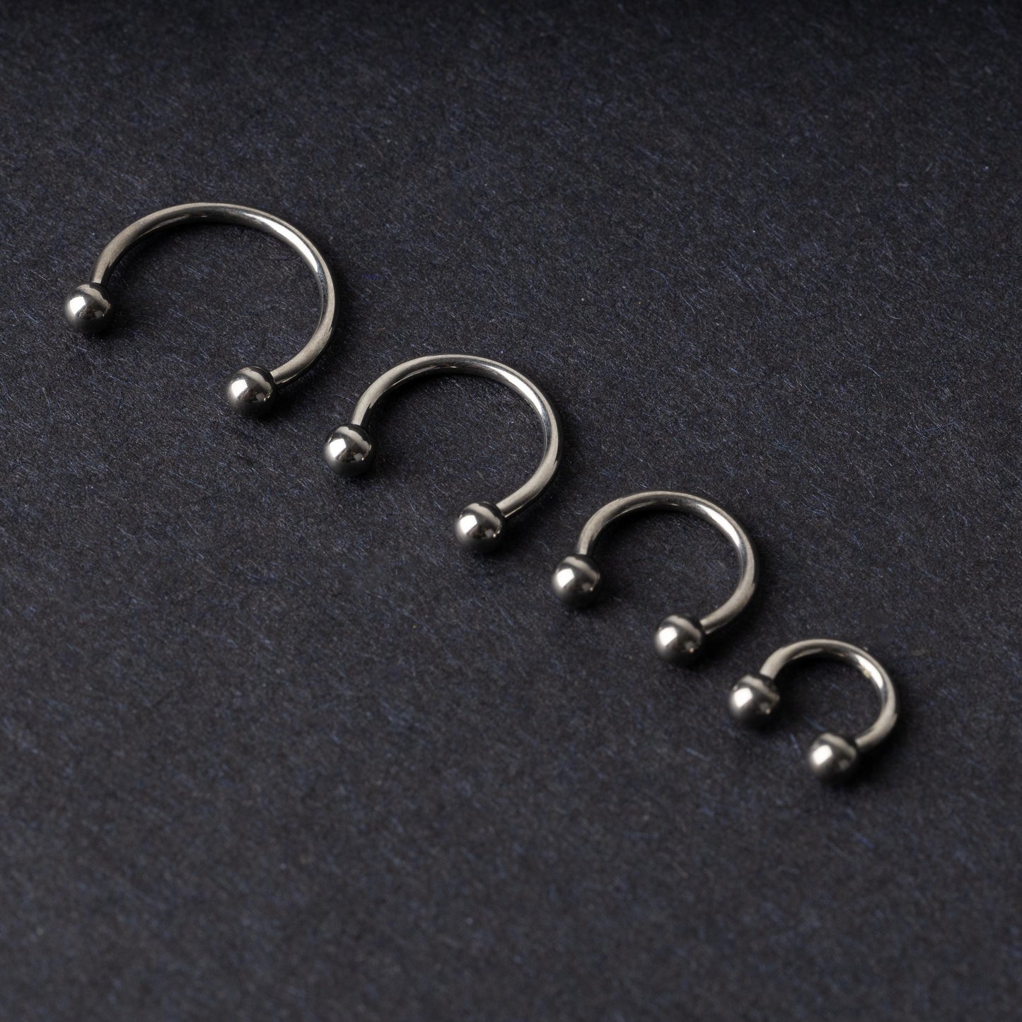 Four titanium horseshoe hoops with ball ends, arranged from largest to smallest on a dark background.