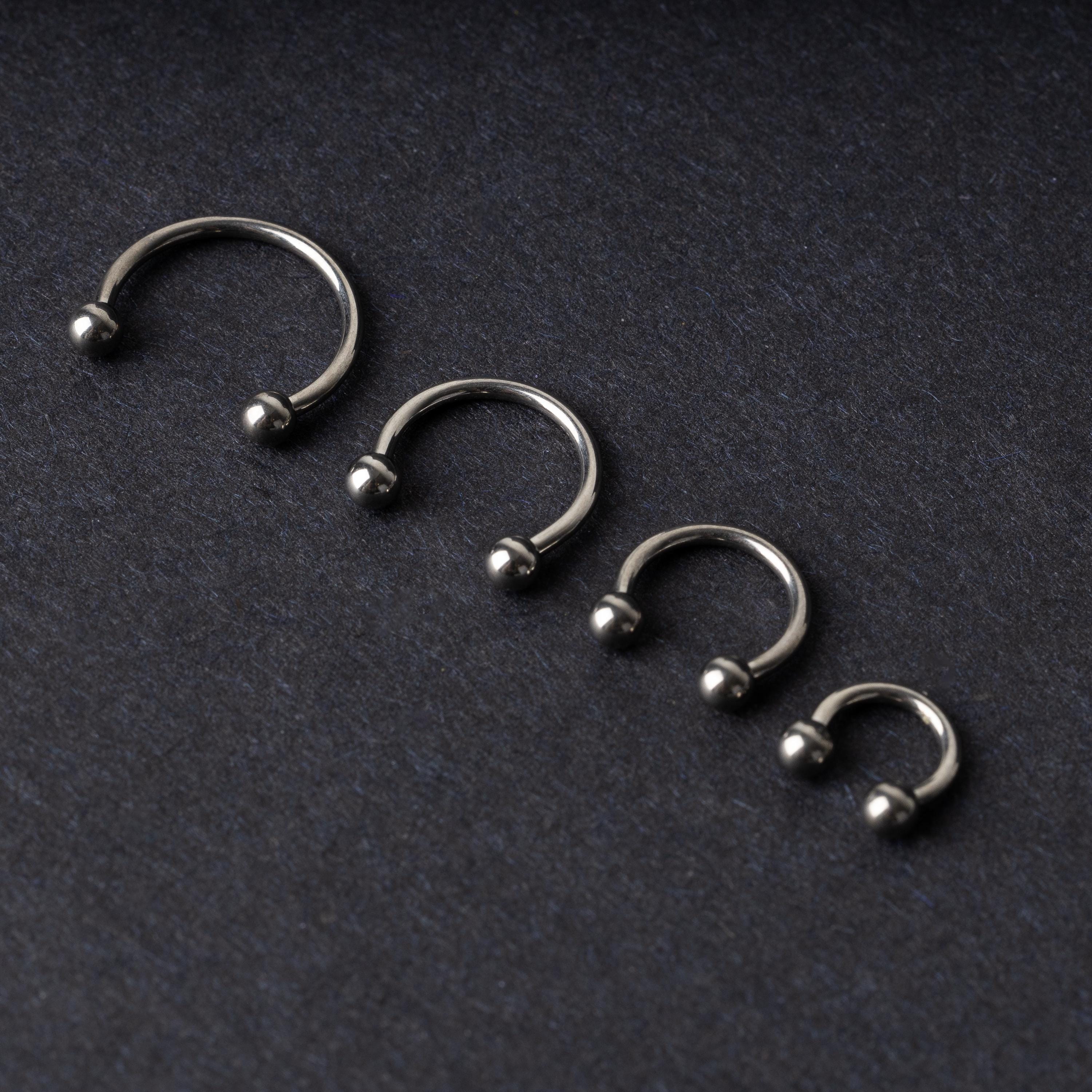 Four titanium horseshoe hoops with ball ends, arranged from largest to smallest on a dark background.