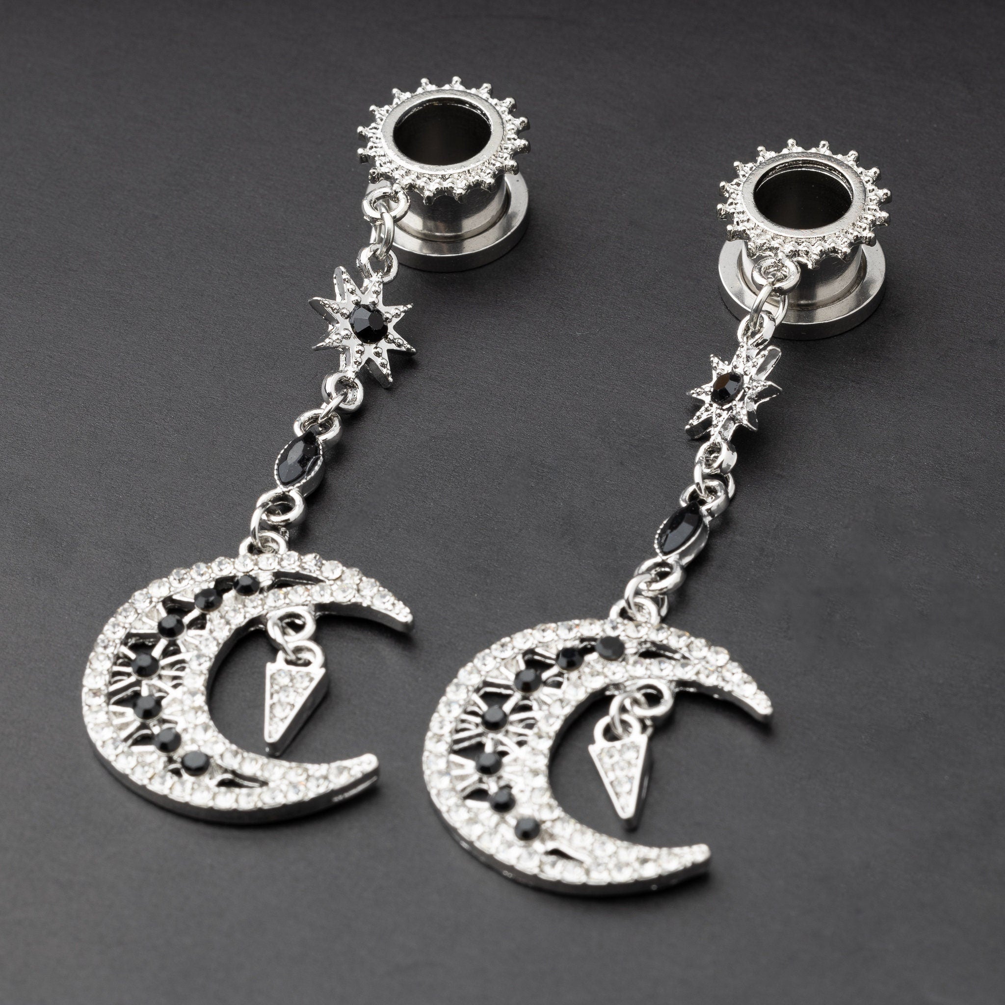 A pair of silver ear hangers for stretched ears with dangling crescent moon and star designs, adorned with black and clear rhinestones, displayed on a dark background.
