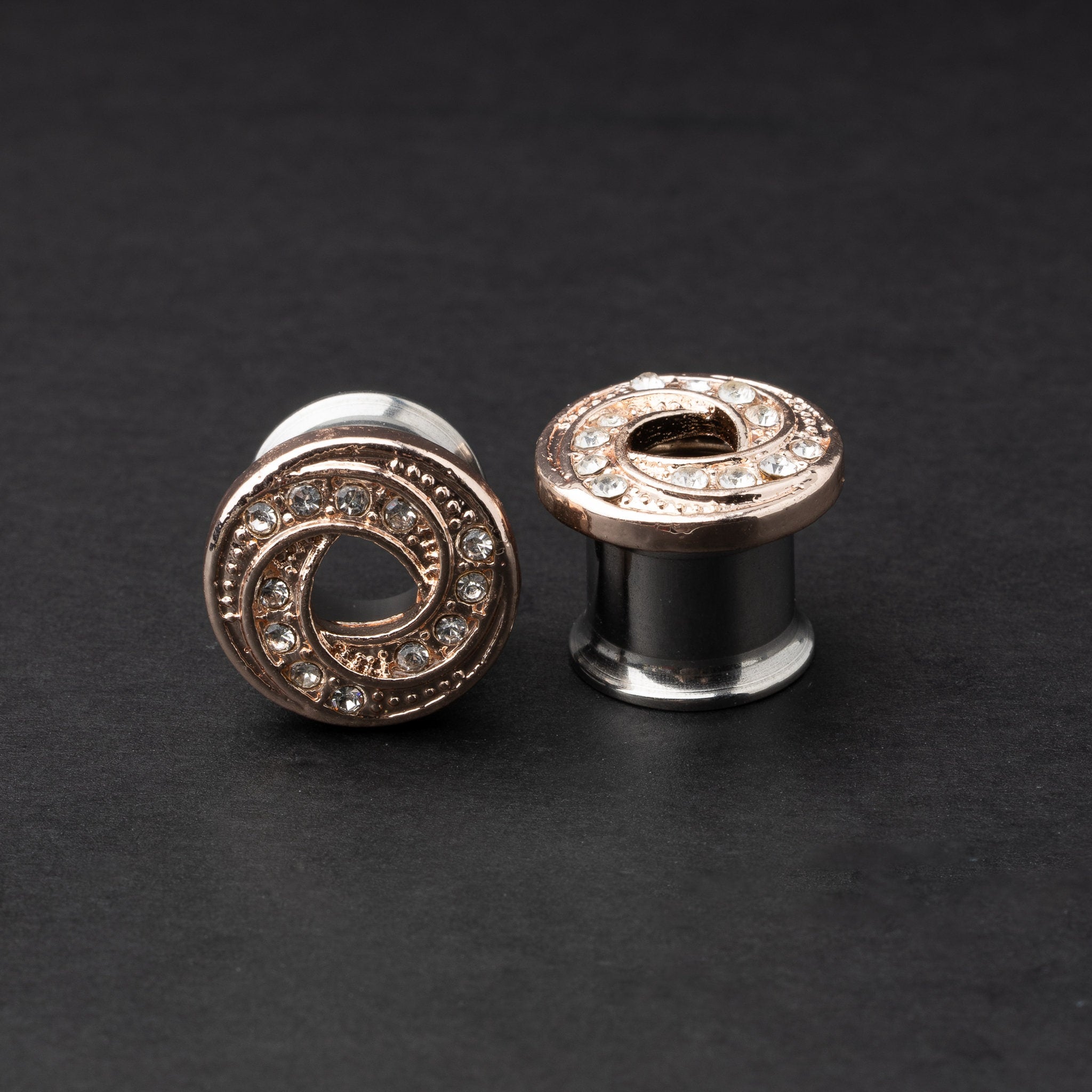 A pair of rose gold-tone stainless steel screw-fit tunnels with an infinity swirl design encrusted with clear crystals, displayed against a dark background.