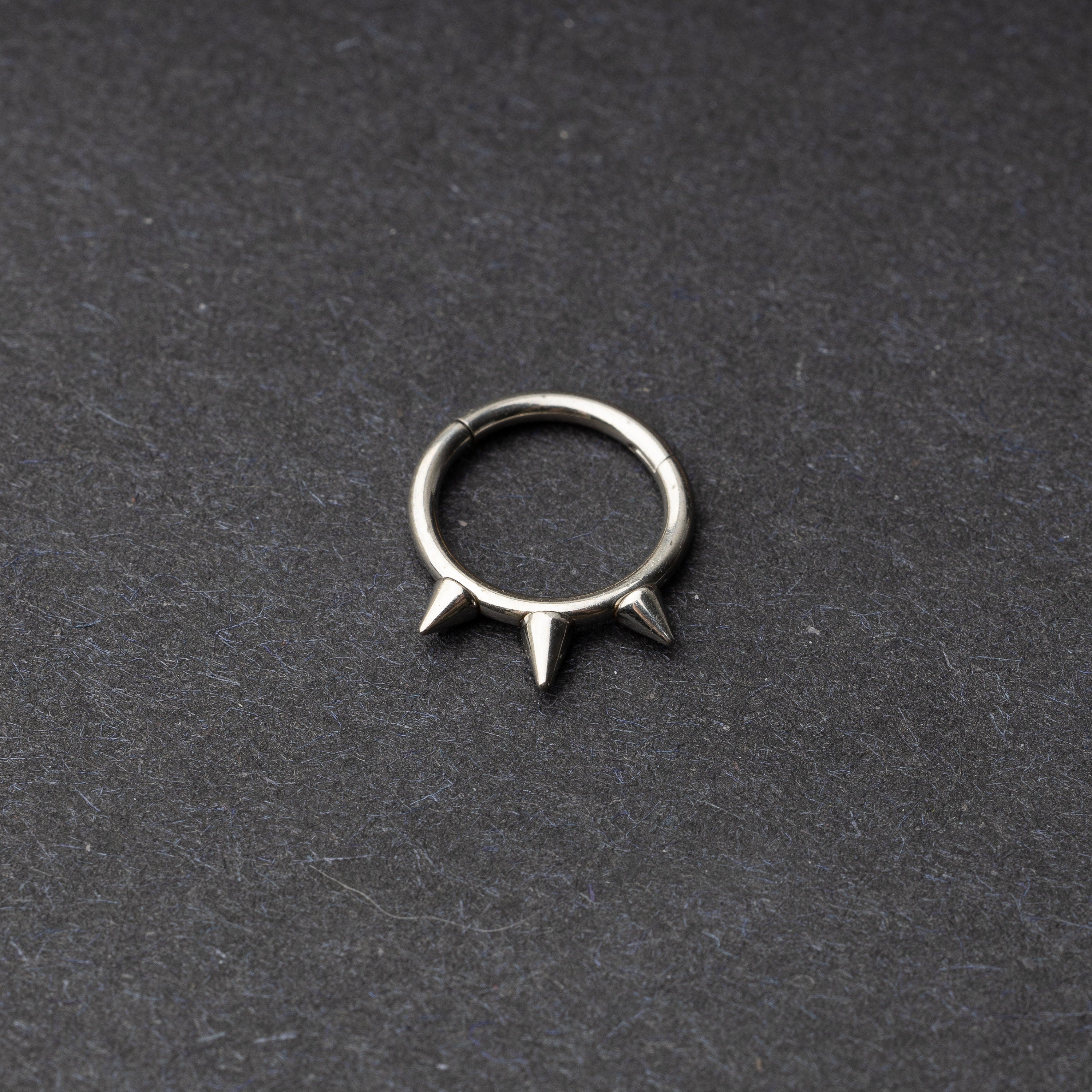 Silver-tone stainless steel clicker ring with three spikes, displayed on a dark textured background.