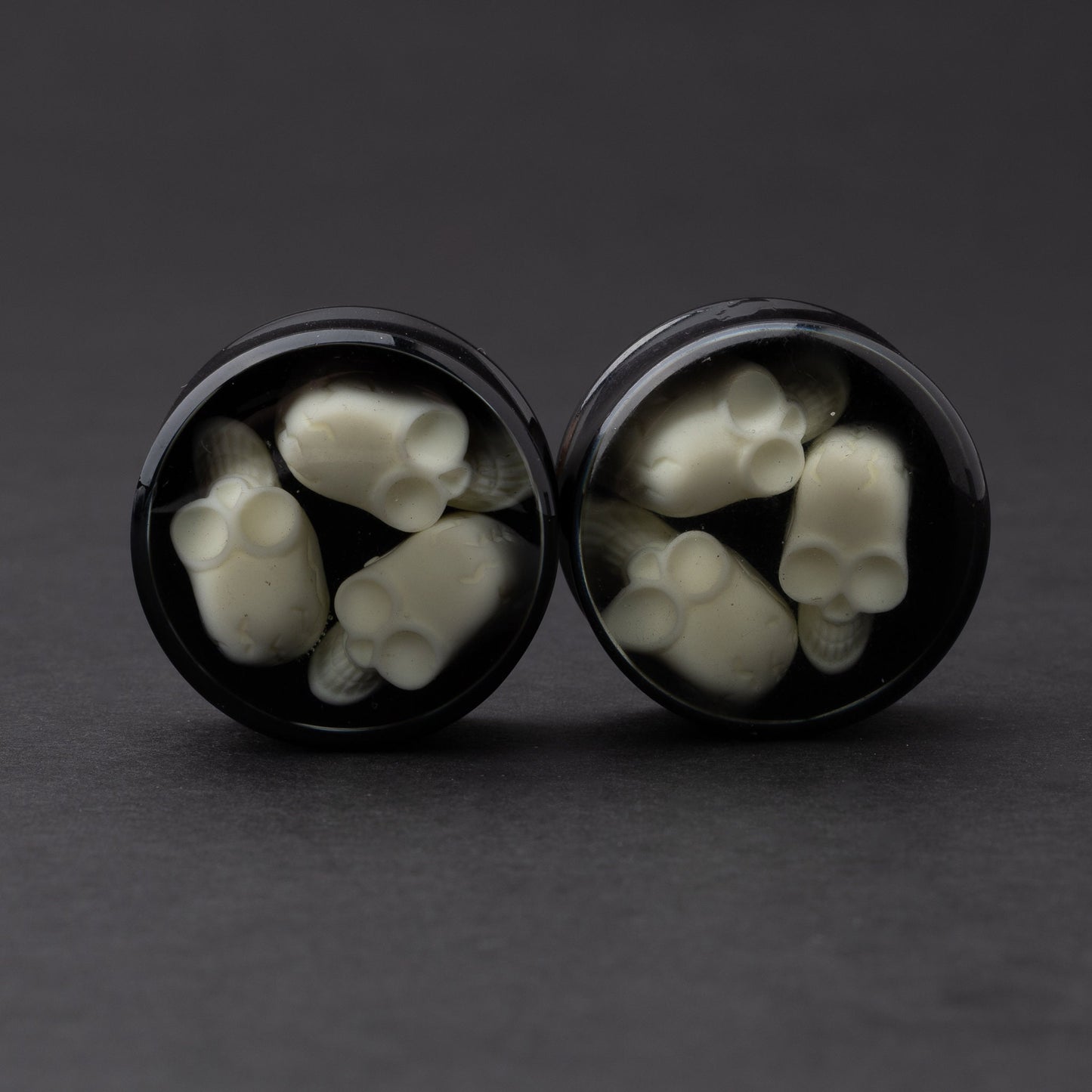 A pair of black double-flared plugs with glow-in-the-dark skulls encased in a polished finish, displayed on a dark background.