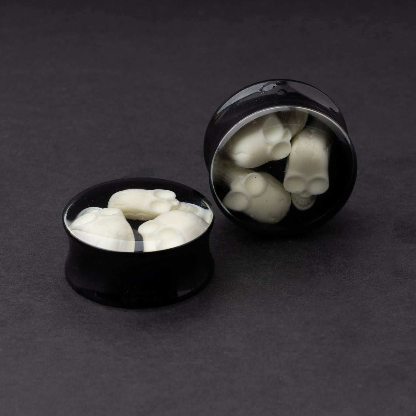 A pair of black double-flared plugs with glow-in-the-dark skulls encased in a polished finish, displayed on a dark background.