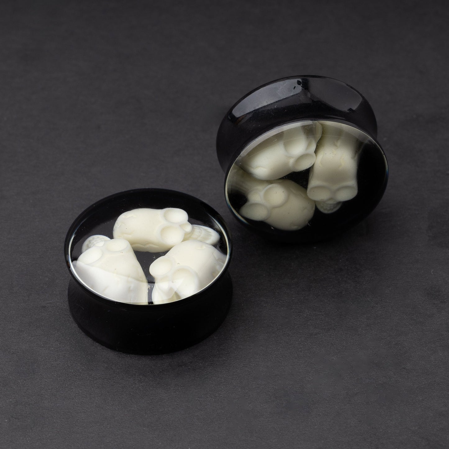A pair of black double-flared plugs with glow-in-the-dark skulls encased in a polished finish, displayed on a dark background.