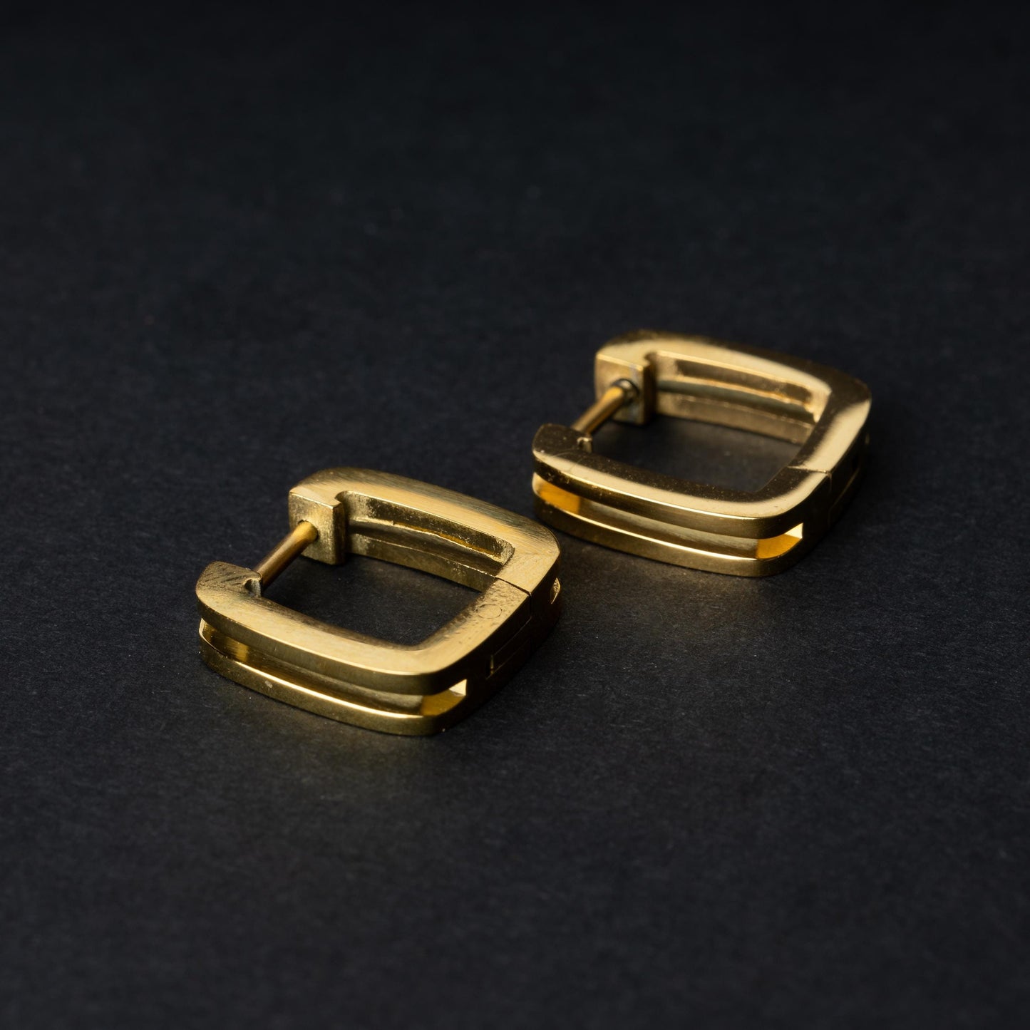 A pair of square-shaped gold hoop earrings displayed on a dark background.