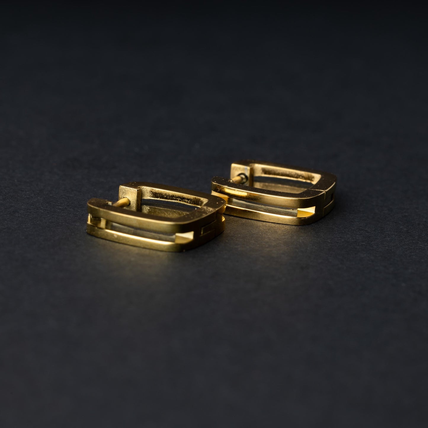 A pair of square-shaped gold hoop earrings displayed on a dark background.
