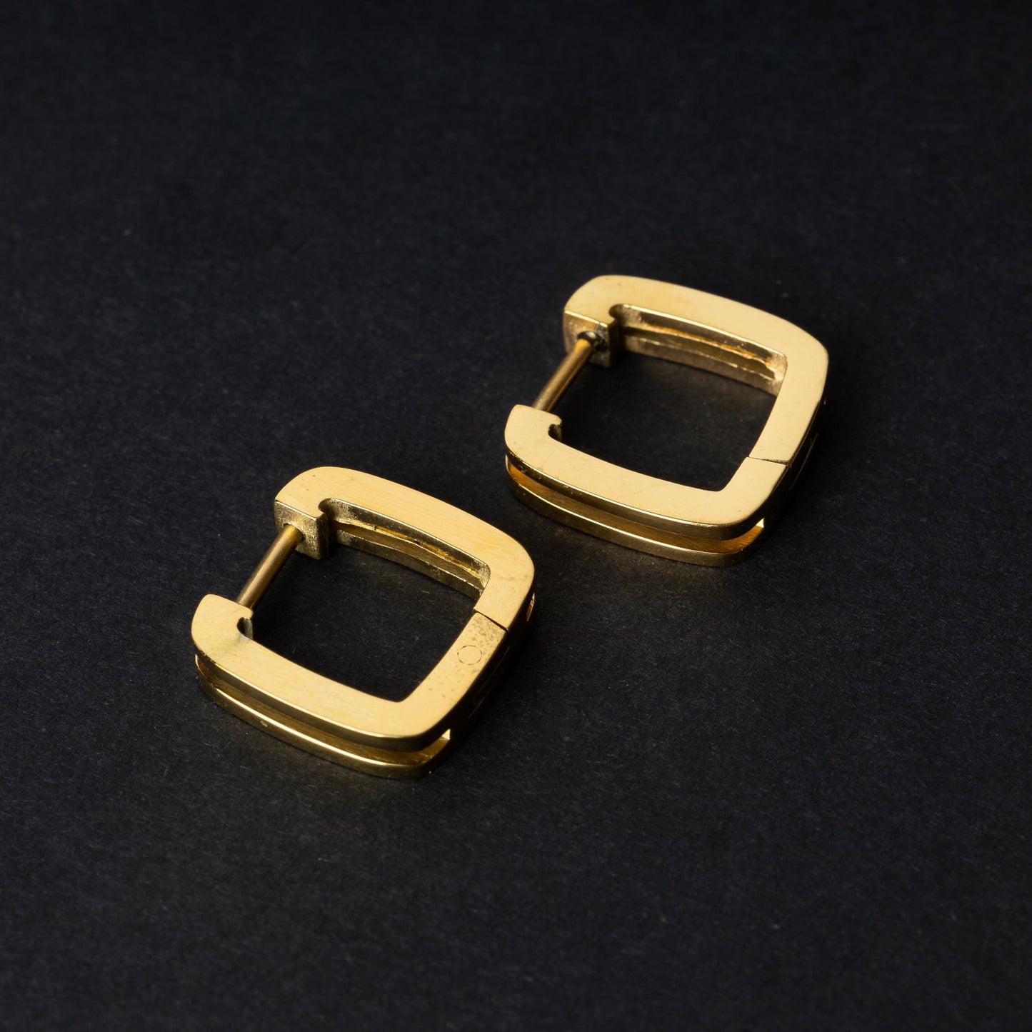 A pair of square-shaped gold hoop earrings displayed on a dark background.