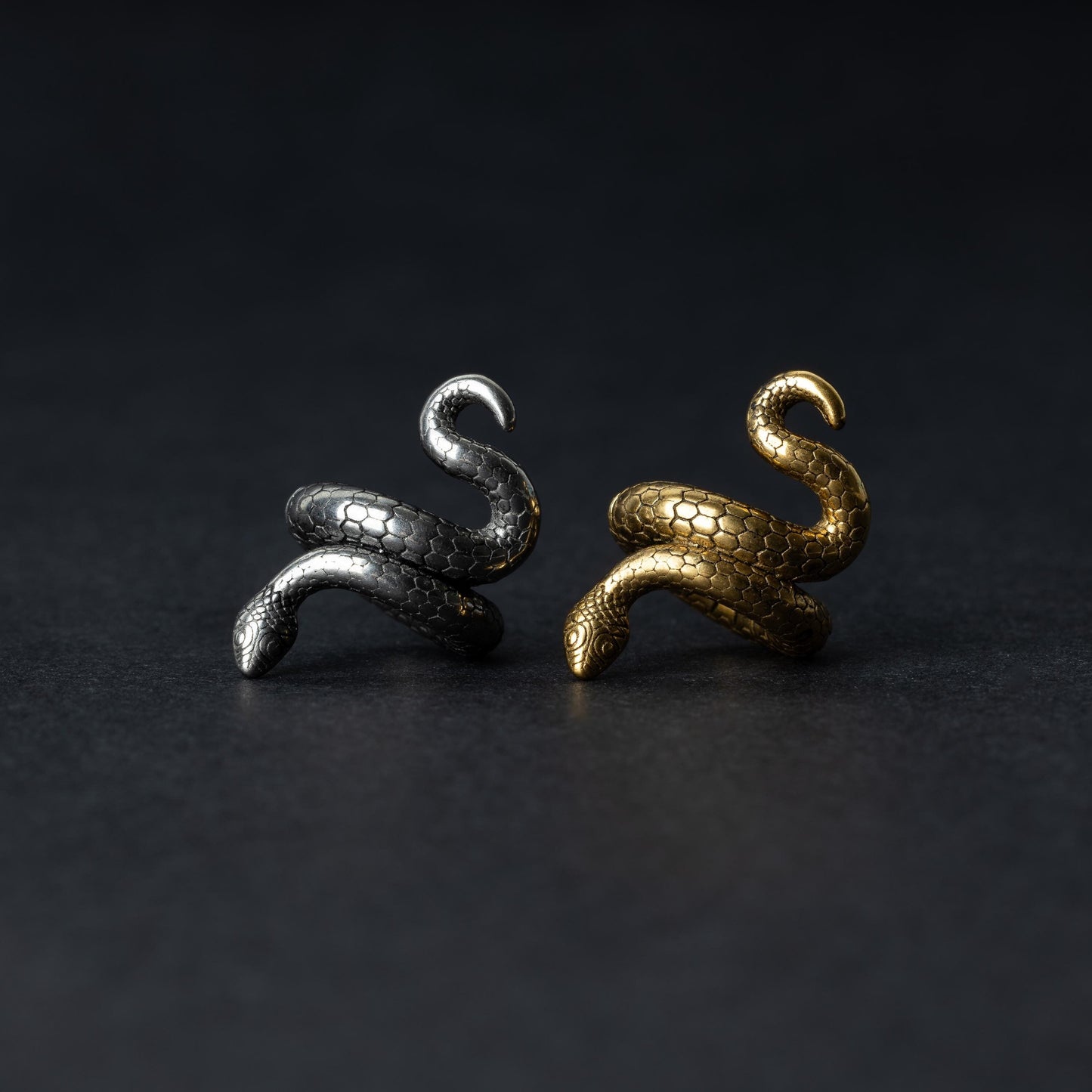 A pair of snake-shaped ear cuffs in polished silver and gold finishes, coiled to wrap around the earlobe, displayed on a dark background.