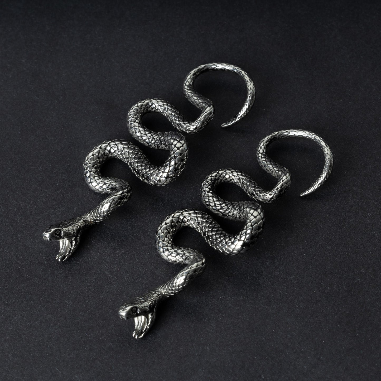 A pair of silver snake-shaped ear weights with intricate scale detailing, coiled design, and open mouths with visible fangs, displayed on a dark background.