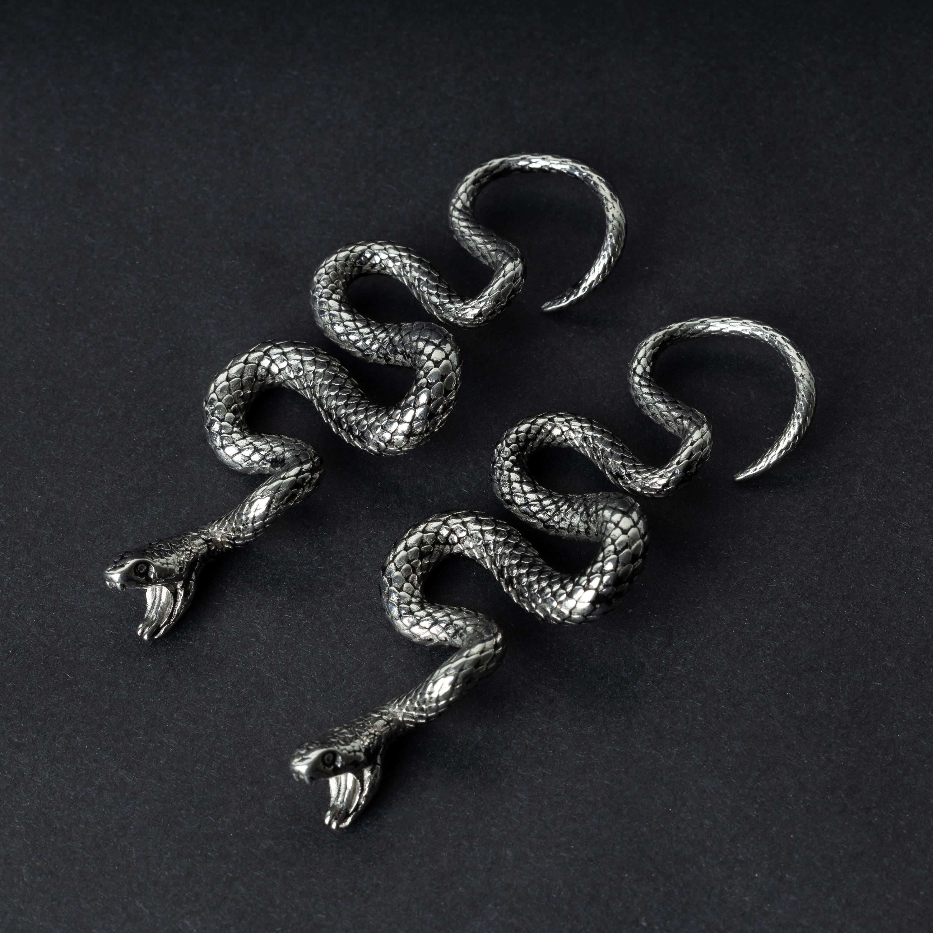 A pair of silver snake-shaped ear weights with intricate scale detailing, coiled design, and open mouths with visible fangs, displayed on a dark background.
