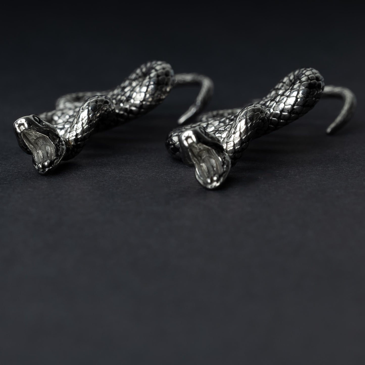 A pair of silver snake-shaped ear weights with intricate scale detailing, coiled design, and open mouths with visible fangs, displayed on a dark background.