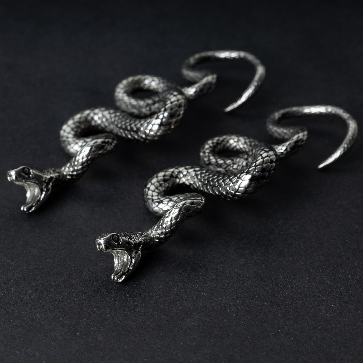 A pair of silver snake-shaped ear weights with intricate scale detailing, coiled design, and open mouths with visible fangs, displayed on a dark background.