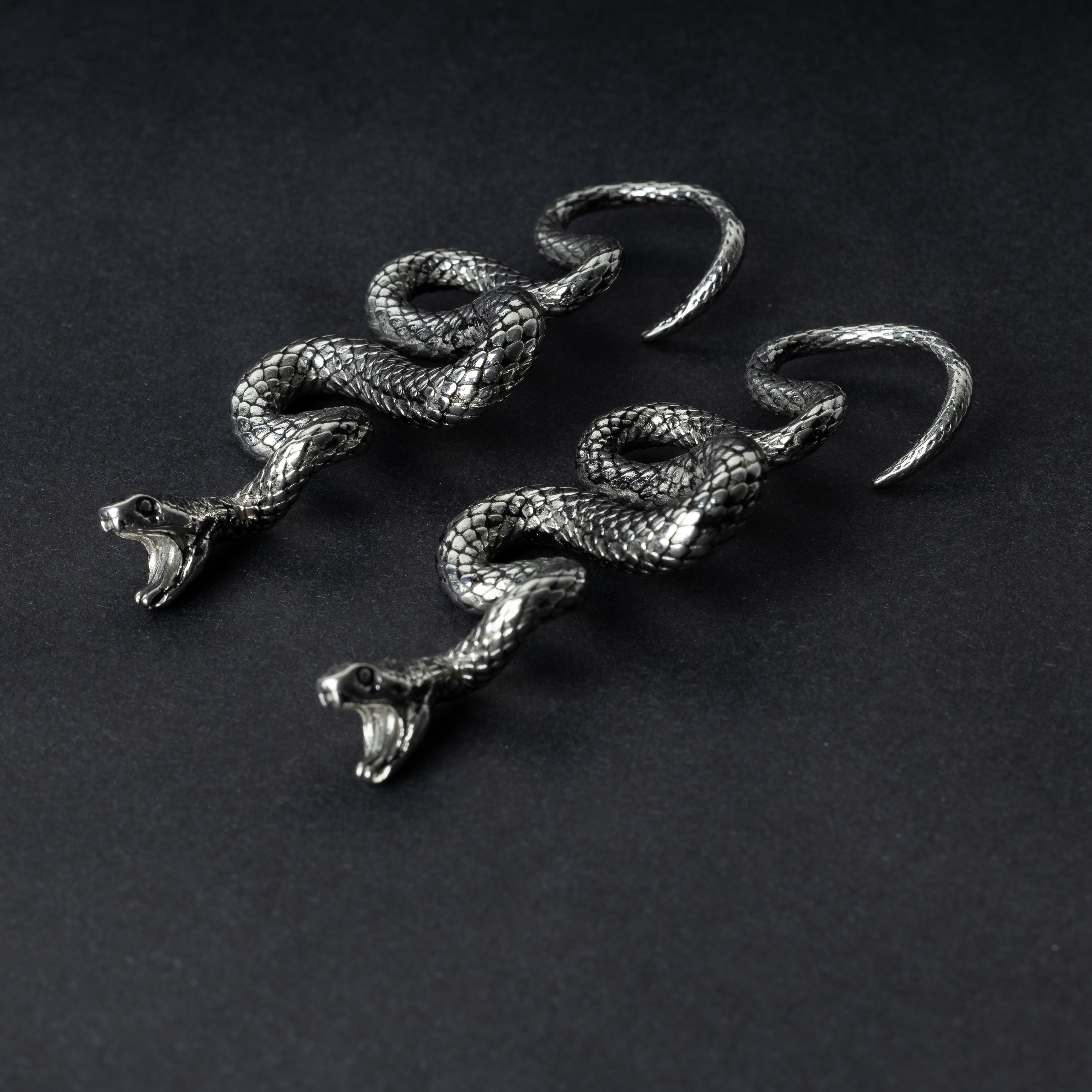 A pair of silver snake-shaped ear weights with intricate scale detailing, coiled design, and open mouths with visible fangs, displayed on a dark background.