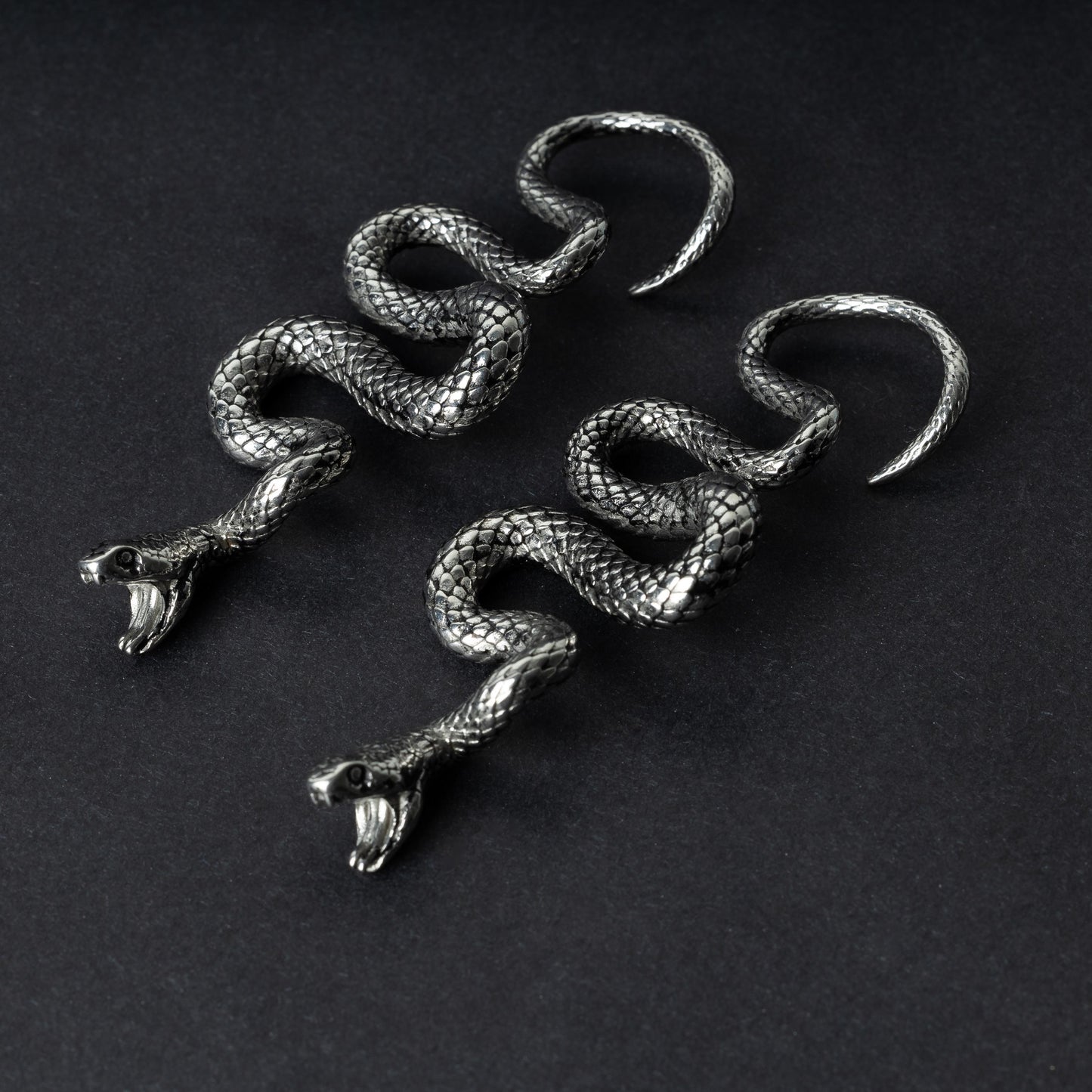 A pair of silver snake-shaped ear weights with intricate scale detailing, coiled design, and open mouths with visible fangs, displayed on a dark background.