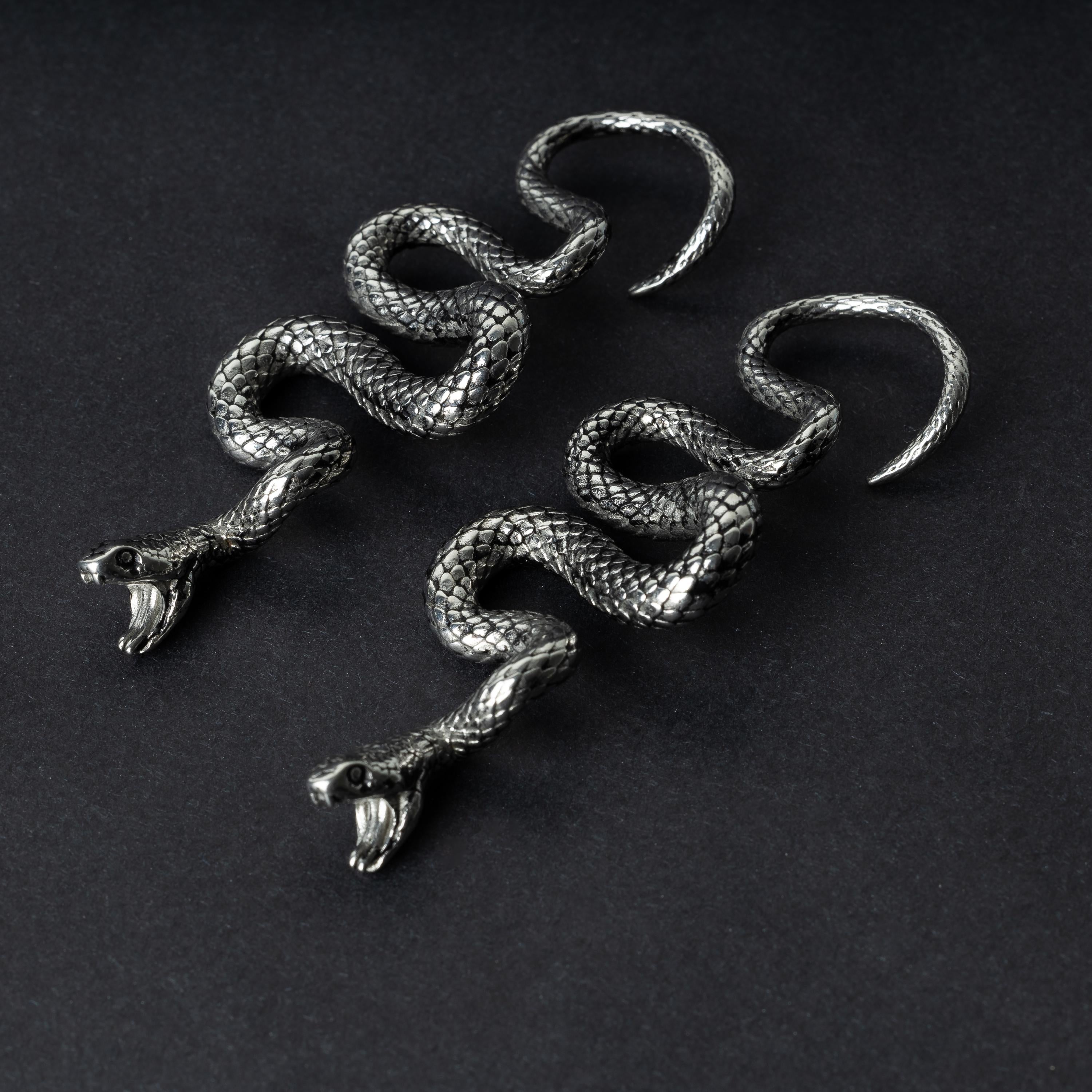 A pair of silver snake-shaped ear weights with intricate scale detailing, coiled design, and open mouths with visible fangs, displayed on a dark background.