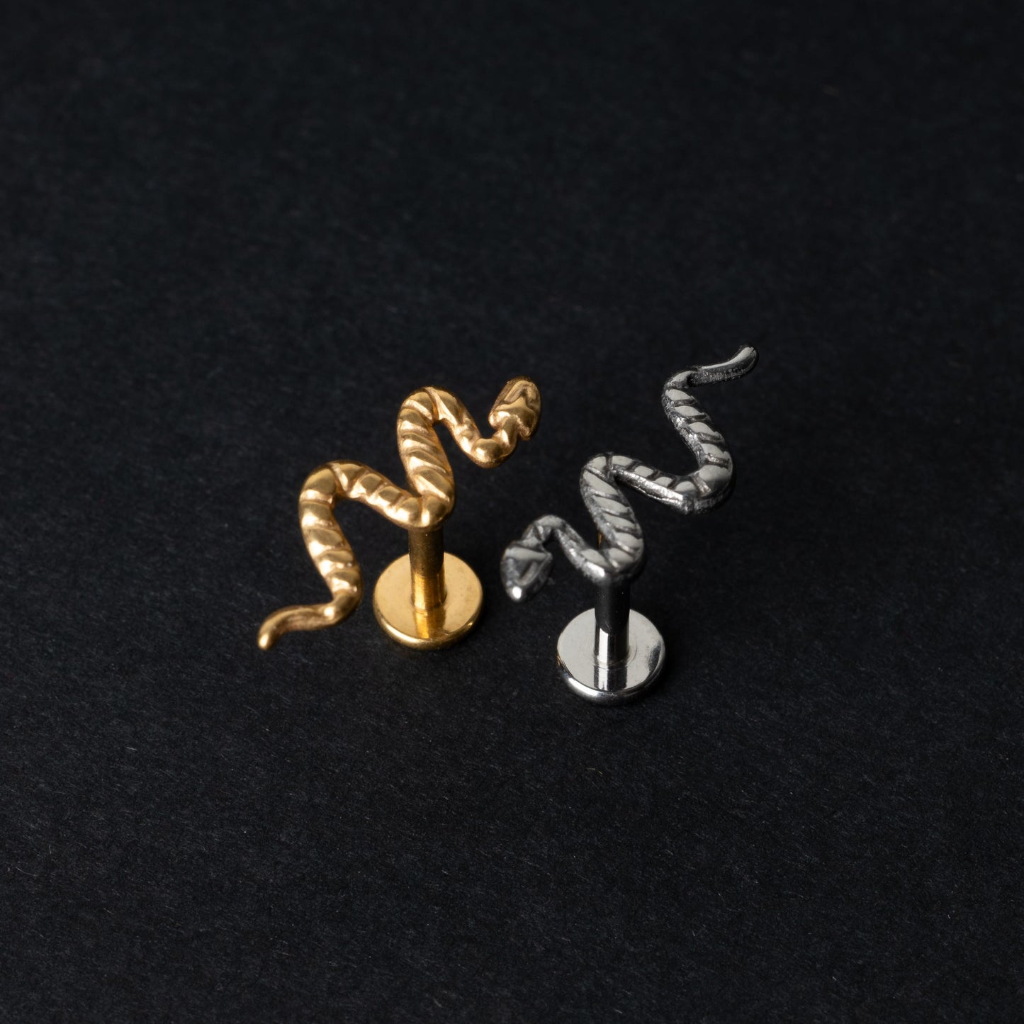 Gold-tone and silver-tone snake labret bars with intricate coiled snake designs displayed on a dark background.
