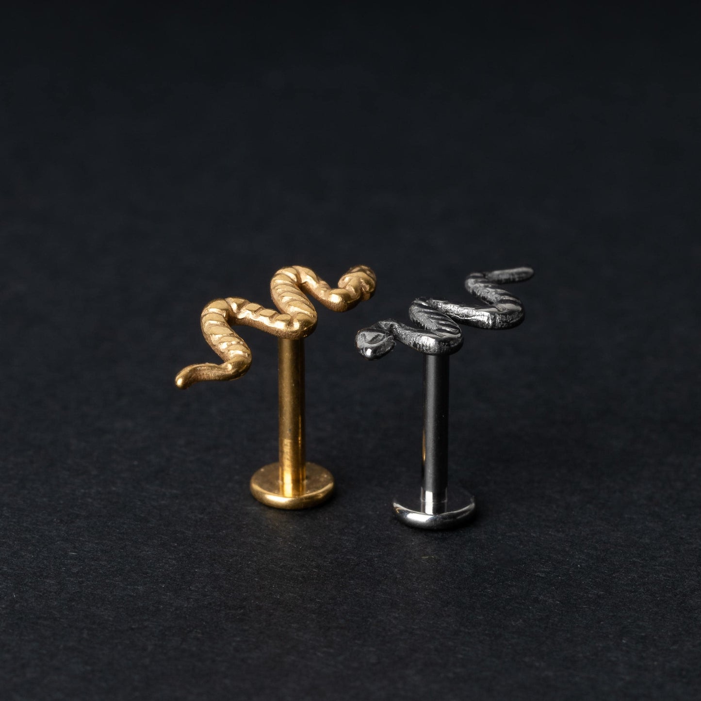 Gold-tone and silver-tone snake labret bars with intricate coiled snake designs displayed on a dark background.