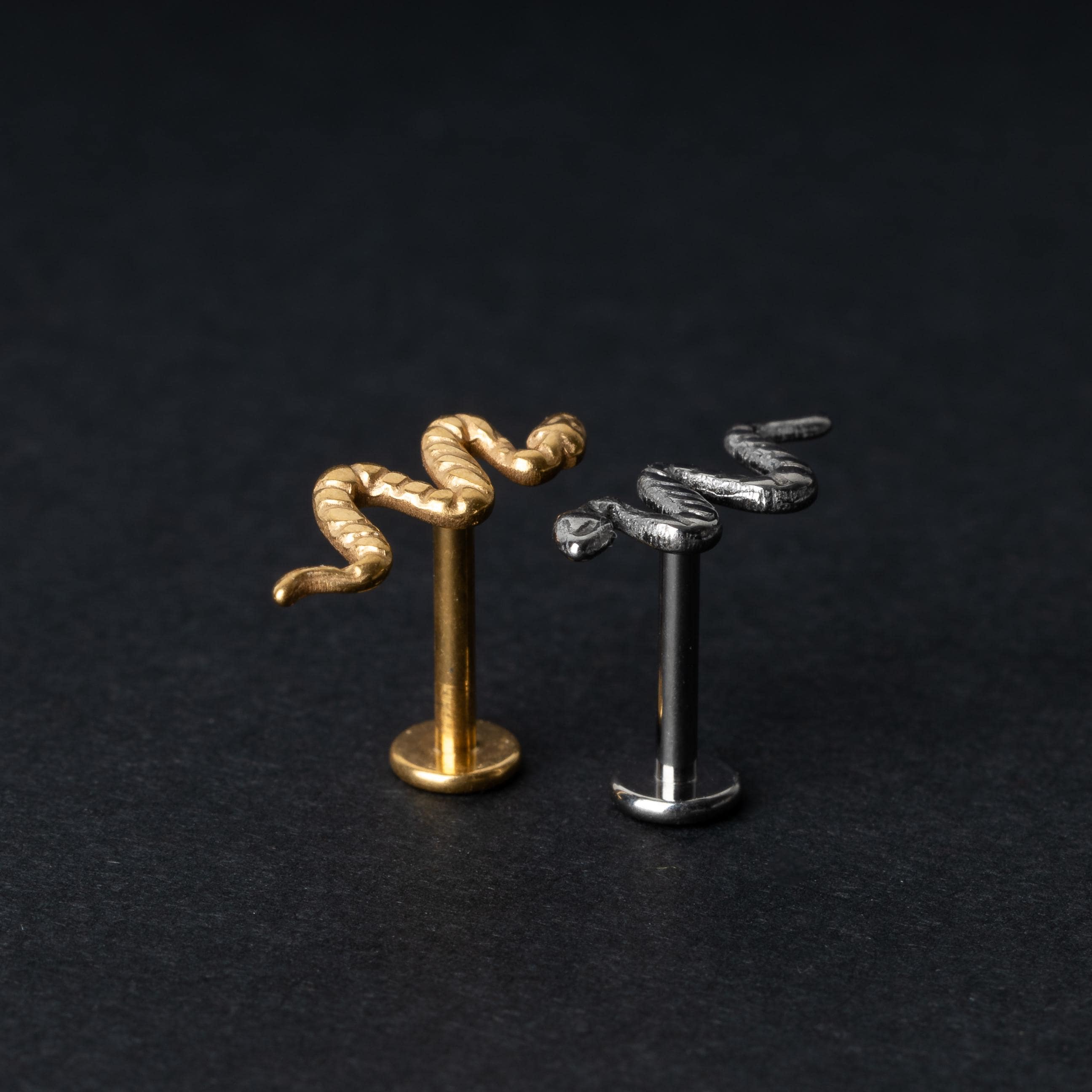 Gold-tone and silver-tone snake labret bars with intricate coiled snake designs displayed on a dark background.