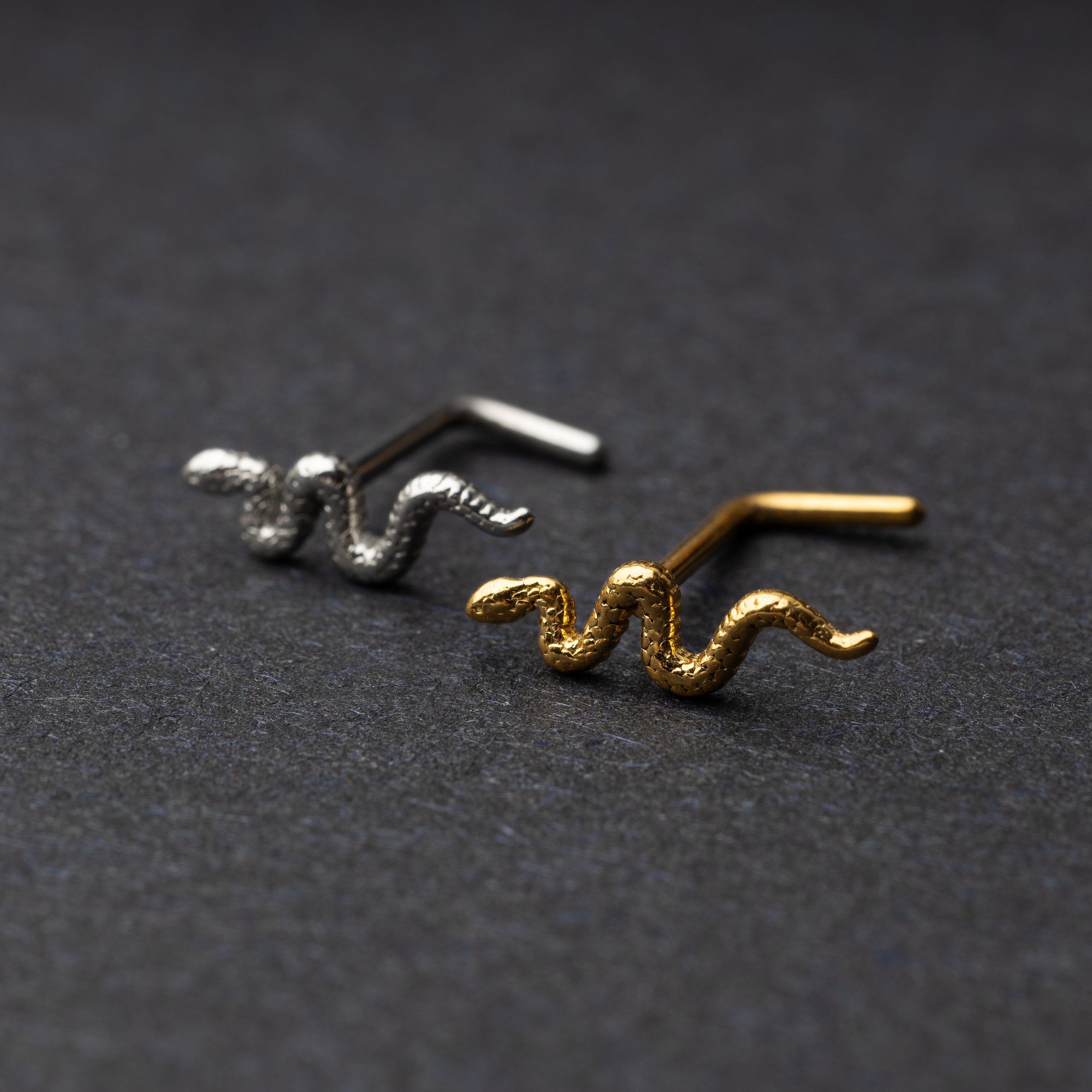 Snake-shaped nose studs in gold and silver finishes displayed on a dark background.