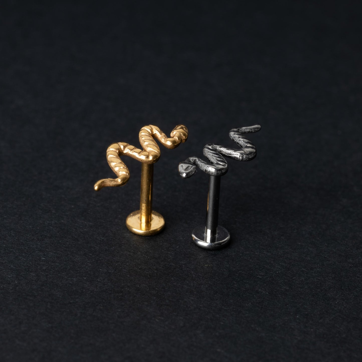 Gold-tone and silver-tone snake labret bars with intricate coiled snake designs displayed on a dark background.