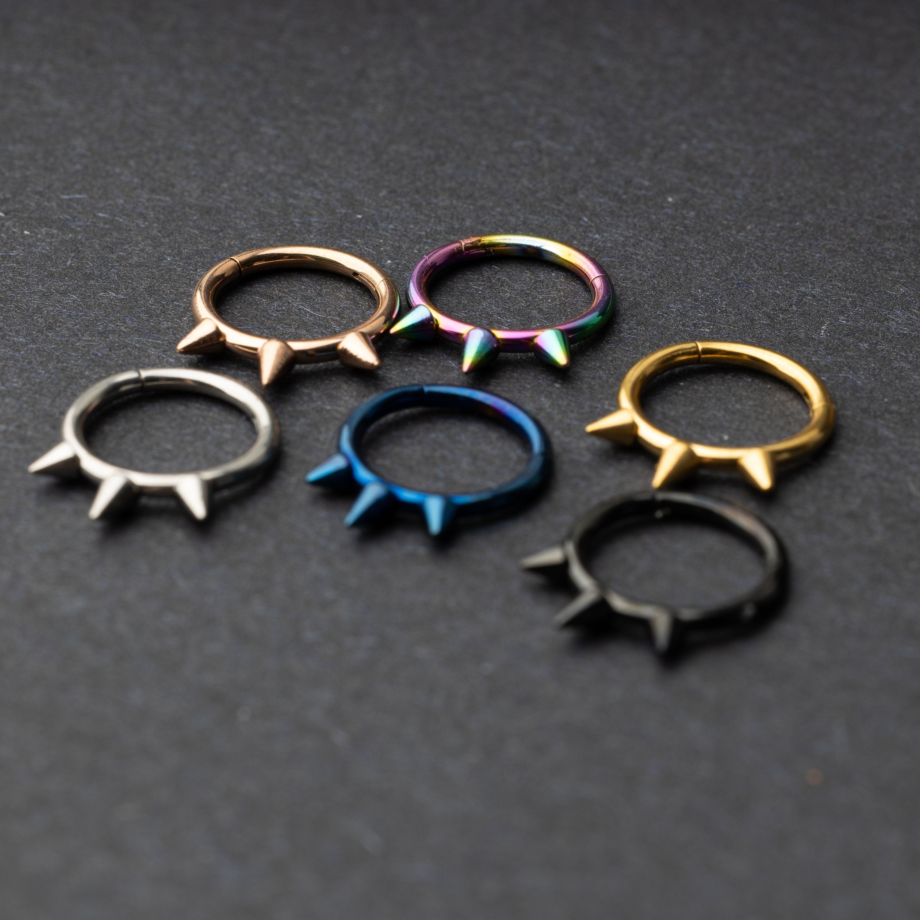Assorted stainless steel spiked rings in various finishes, including gold, black, rainbow, and blue, displayed on a dark textured background.