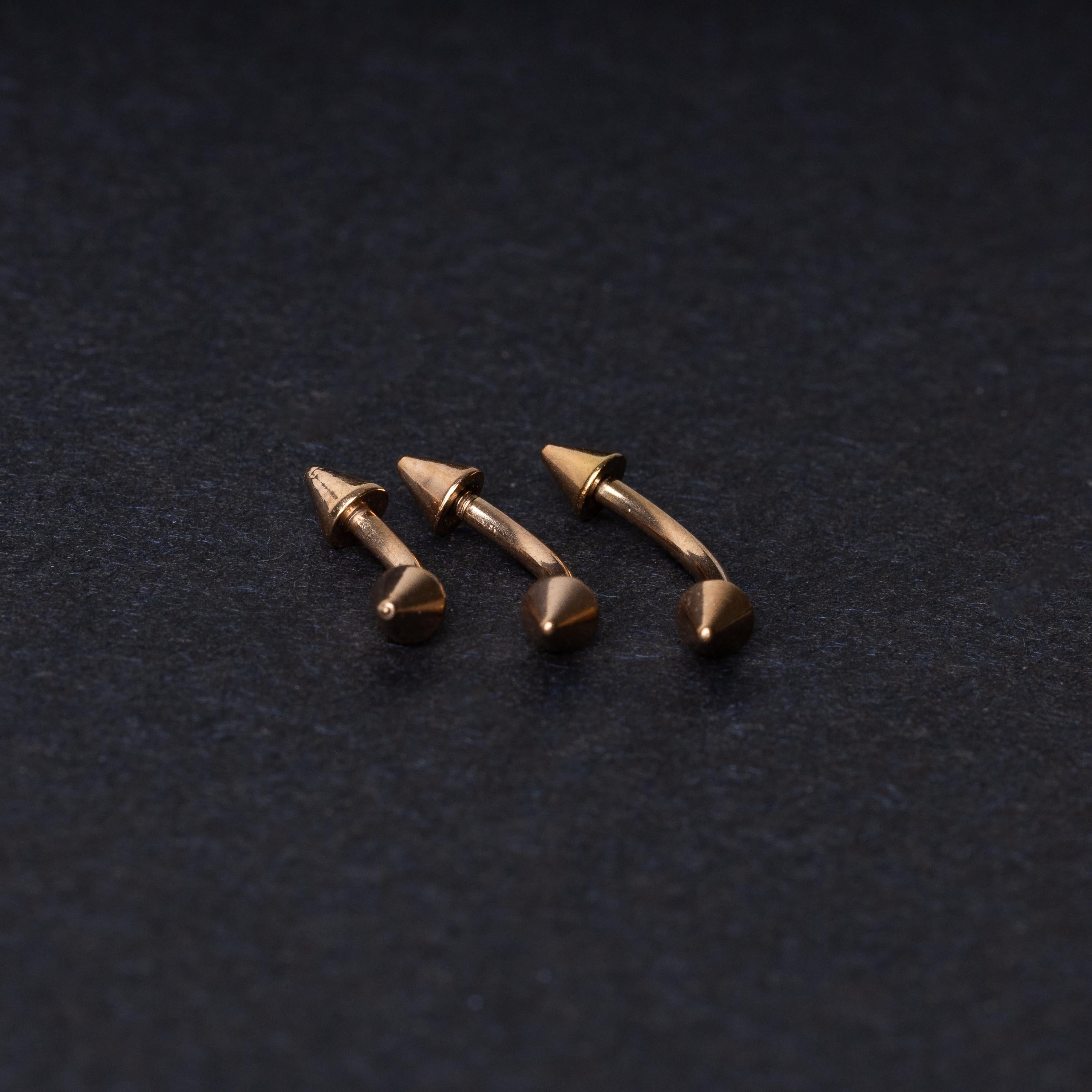 Three gold-tone titanium curved barbells with cone ends, displayed on a dark textured background, showcasing their sleek curved design and polished finish.