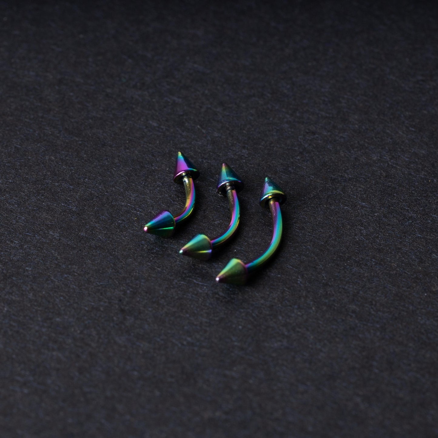 Three rainbow titanium curved barbells with cone-shaped ends, displayed on a dark textured background, showcasing their vivid color gradient and sleek design.