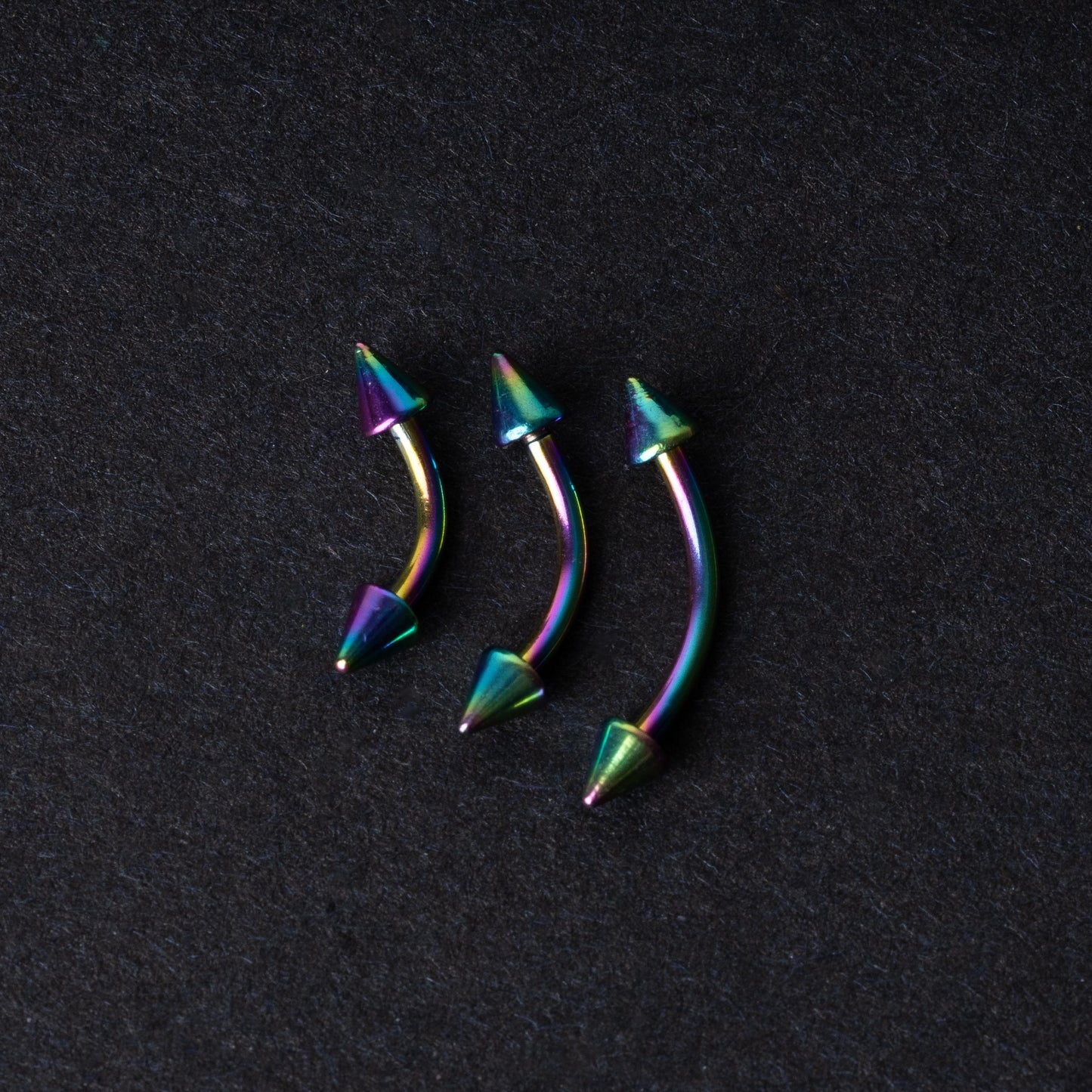 Three rainbow titanium curved barbells with cone-shaped ends, displayed on a dark textured background, showcasing their vivid color gradient and sleek design.