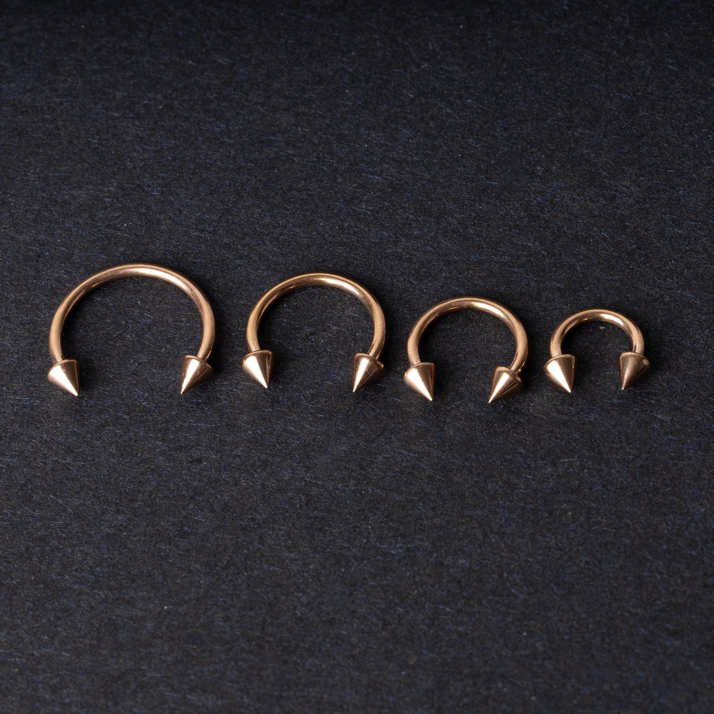 Four rose gold-tone horseshoe hoops with spike ends, arranged in descending size order on a dark textured background.