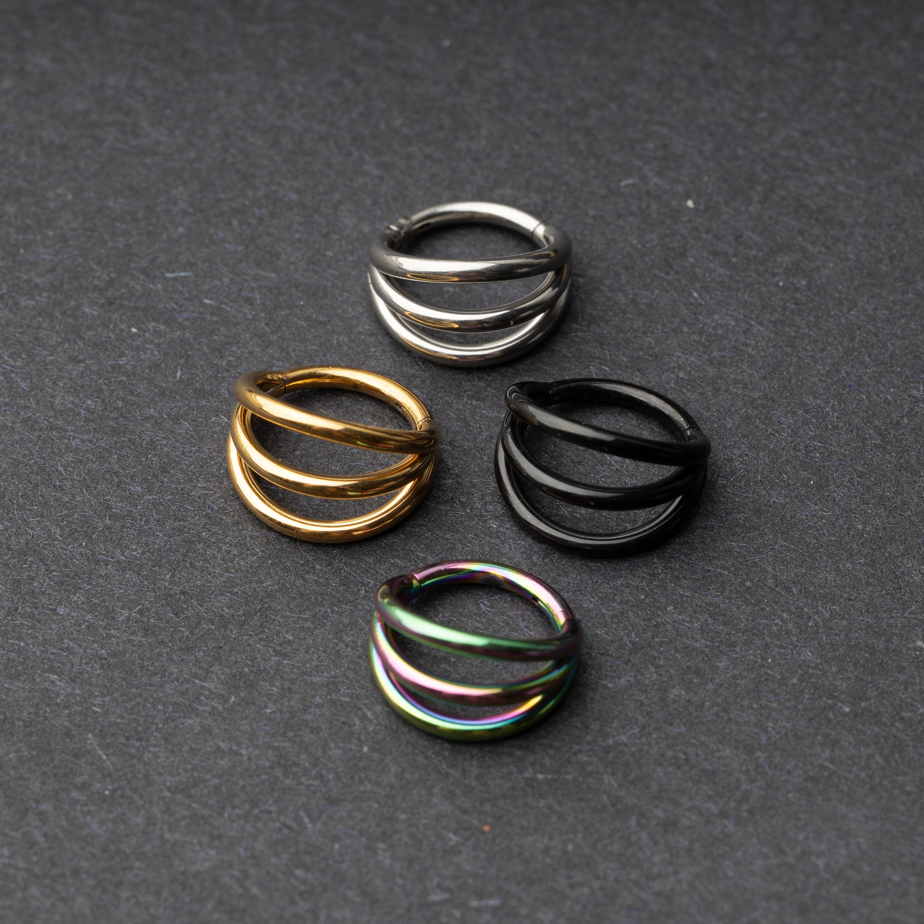 Four triple-band hoop septum clicker rings in gold, silver, black, and rainbow finishes displayed on a dark background.
