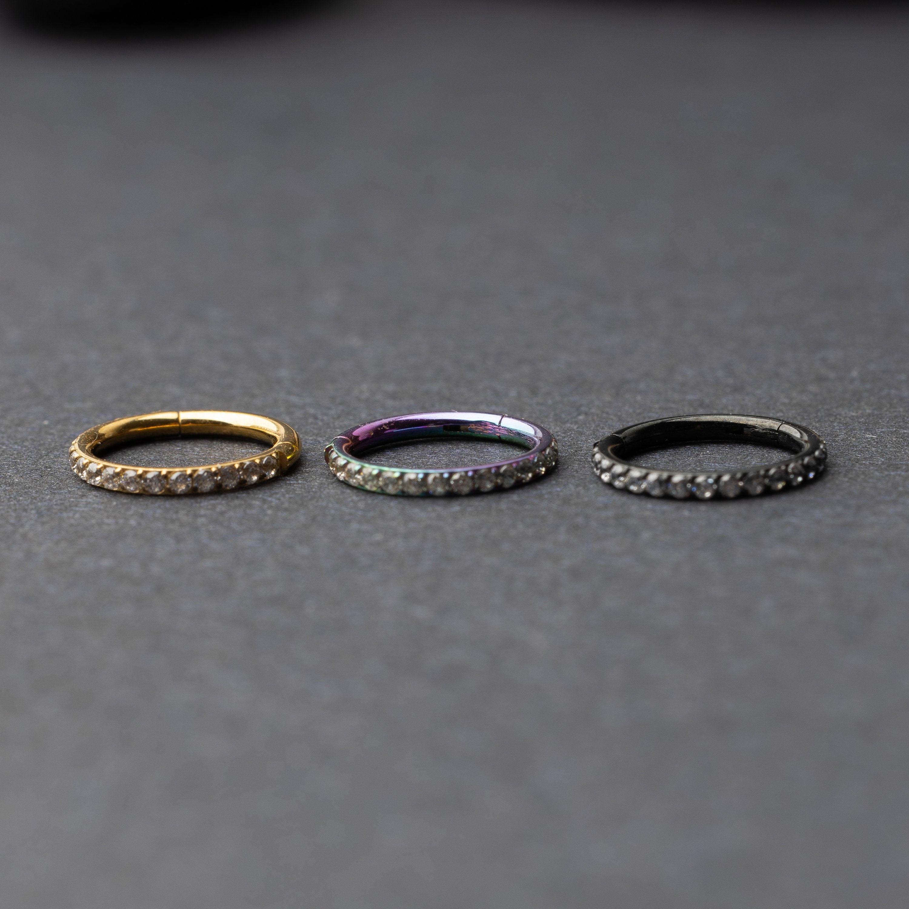 Stainless steel hoops with a row of clear sparkling gems in gold, rainbow and black displayed on a dark background.