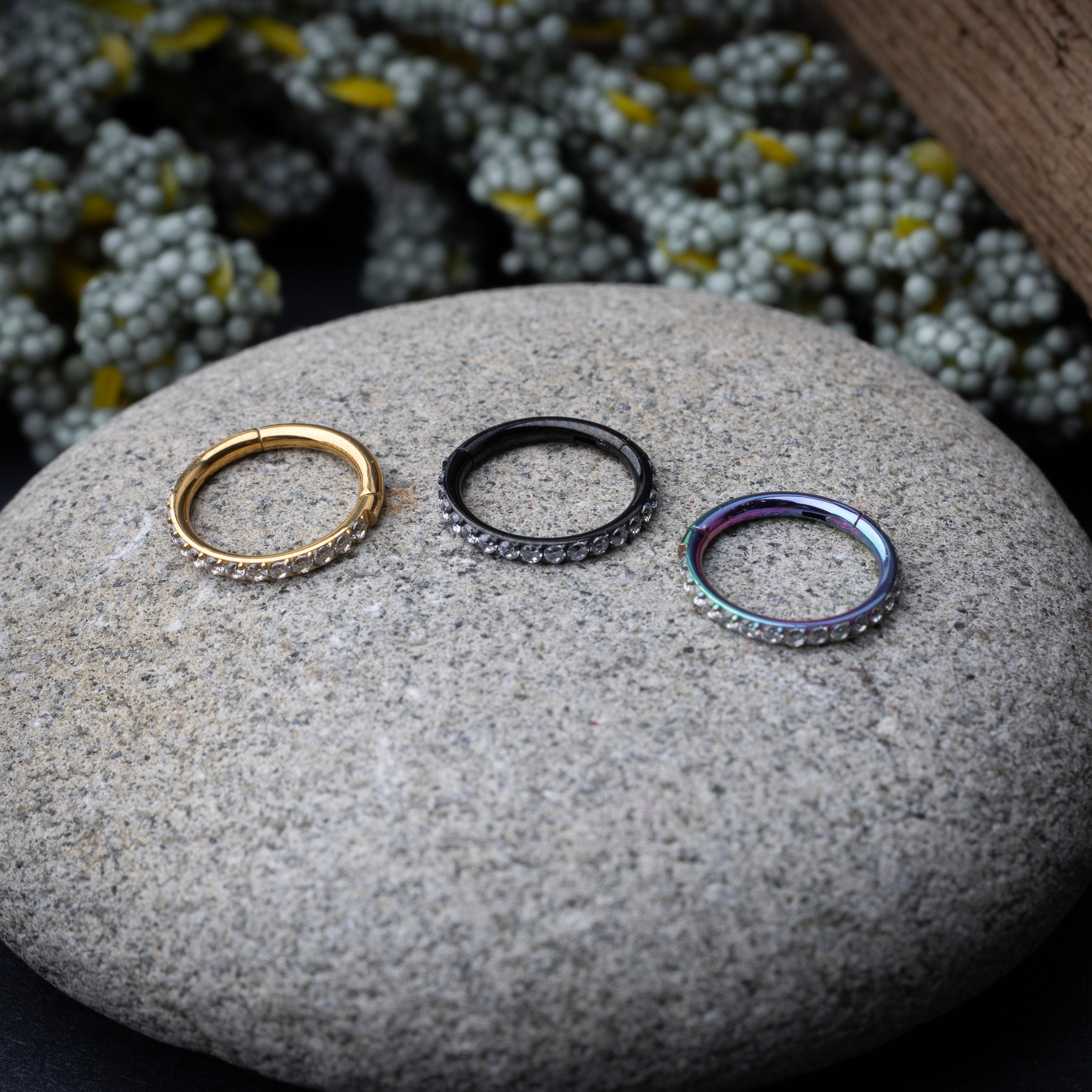 Stainless steel hoops with a row of clear sparkling gems in gold, rainbow and black displayed on a stone background.