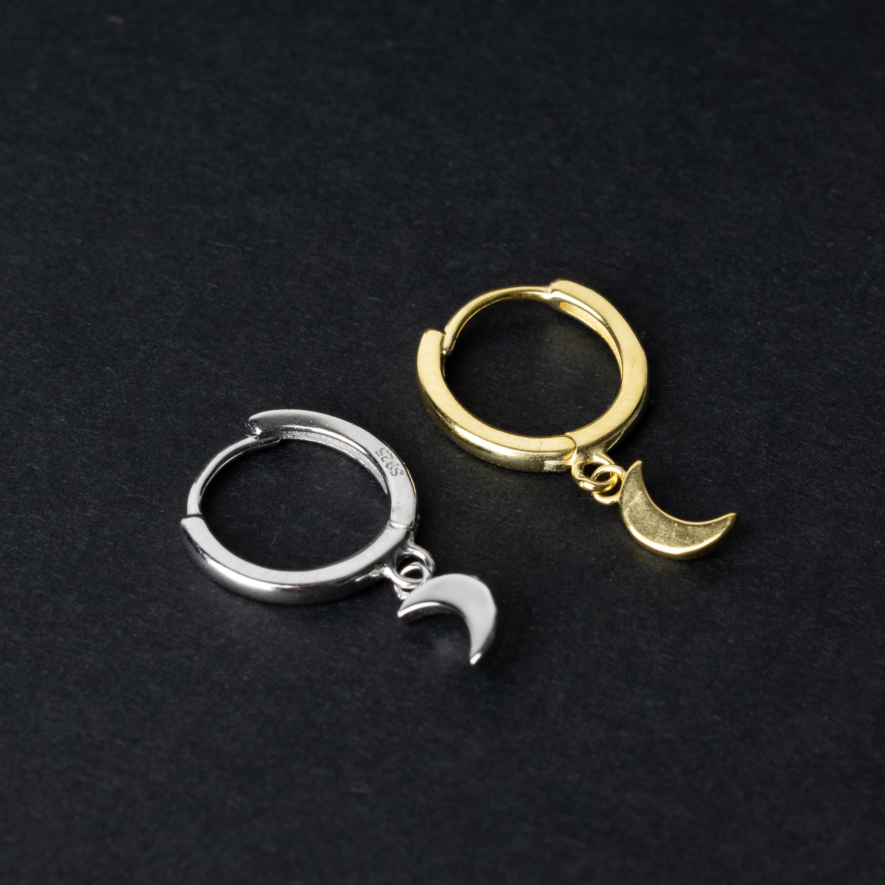 Two crescent moon huggie hoop earrings in gold and silver, each featuring a dangling crescent charm, displayed on a dark background.