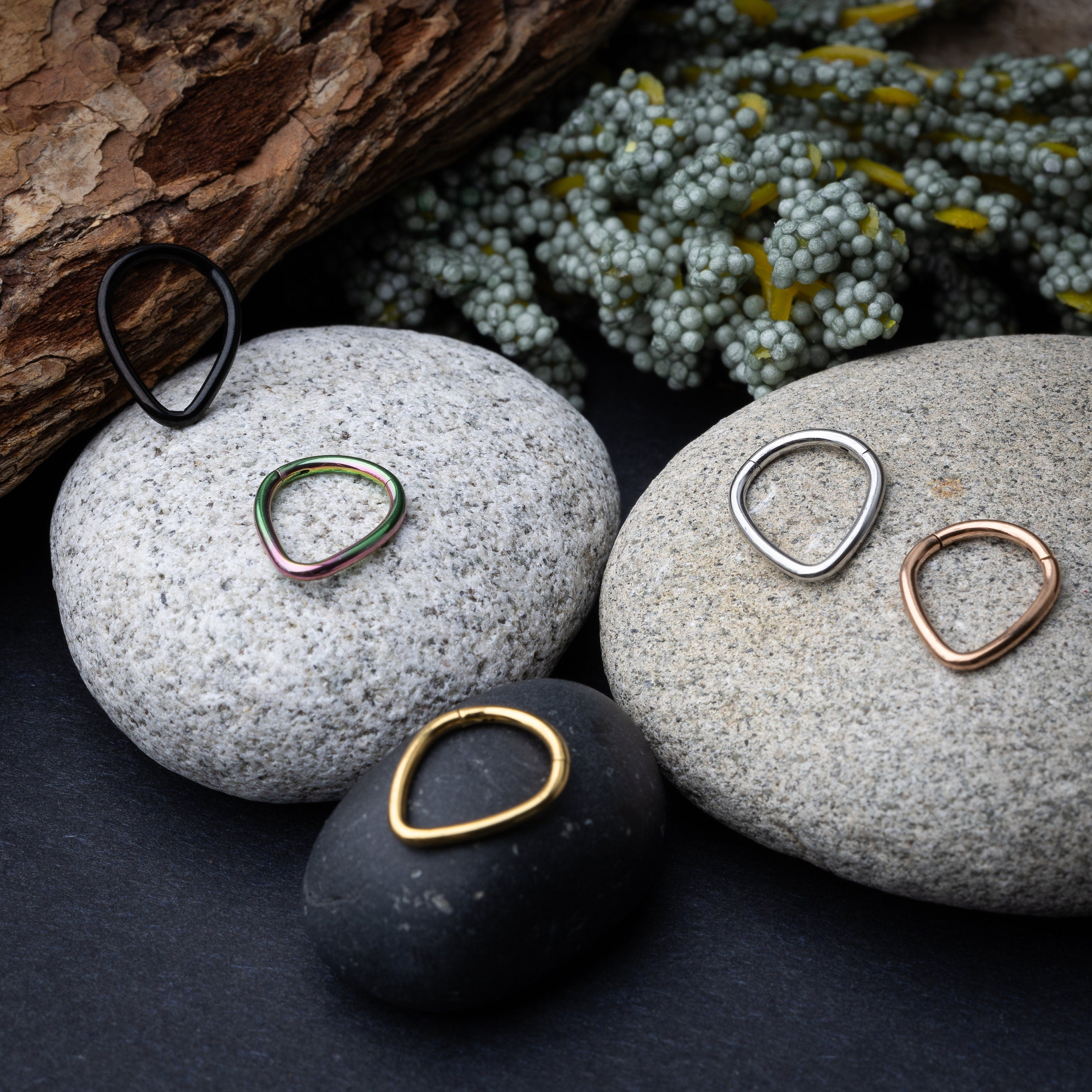 A collection of teardrop-shaped clickers in gold, rainbow, silver, rose gold, and black finishes arranged on a stone and wooden background.