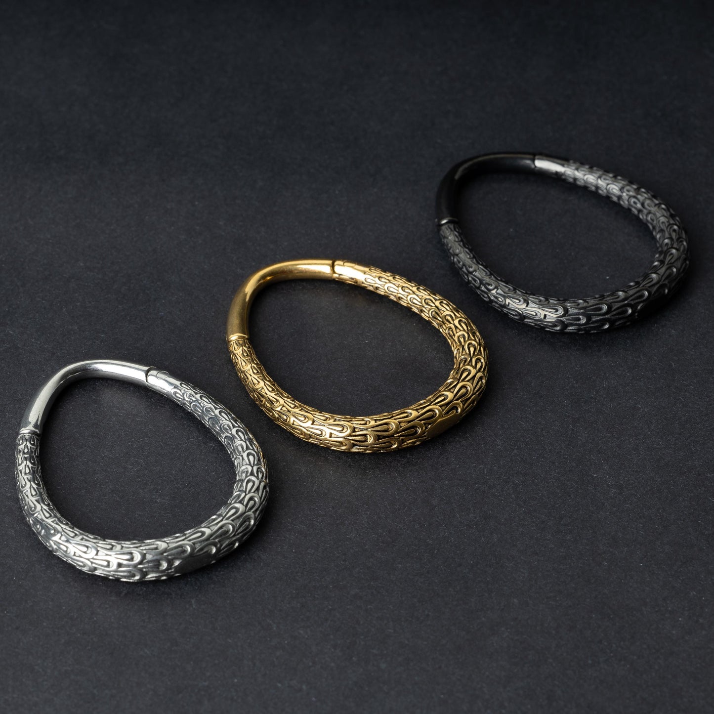 Three teardrop-shaped ear weights with a pebble-textured finish in silver, gold, and black, displayed on a dark background.
