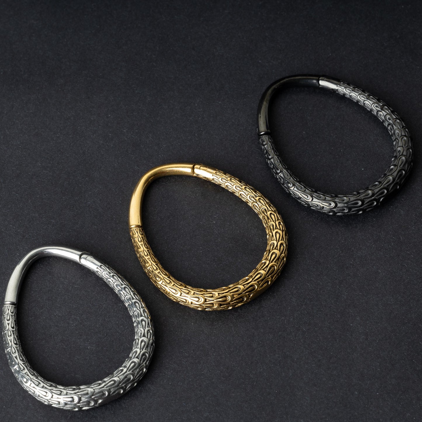 Three teardrop-shaped ear weights with a pebble-textured finish in silver, gold, and black, displayed on a dark background.