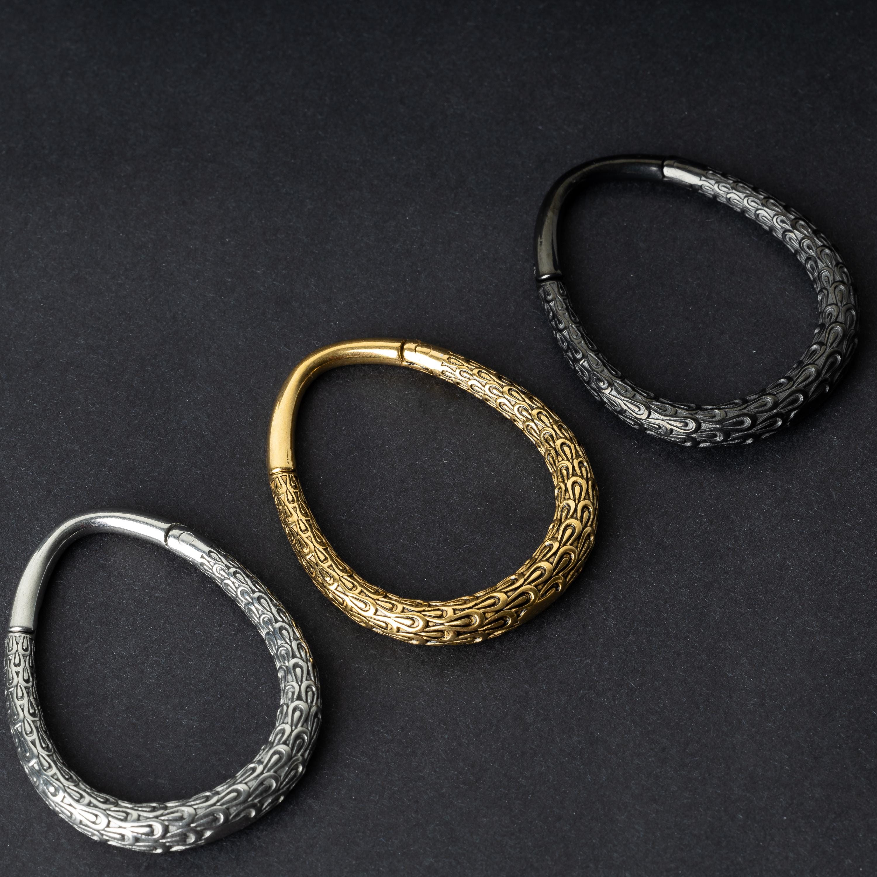 Three teardrop-shaped ear weights with a pebble-textured finish in silver, gold, and black, displayed on a dark background.