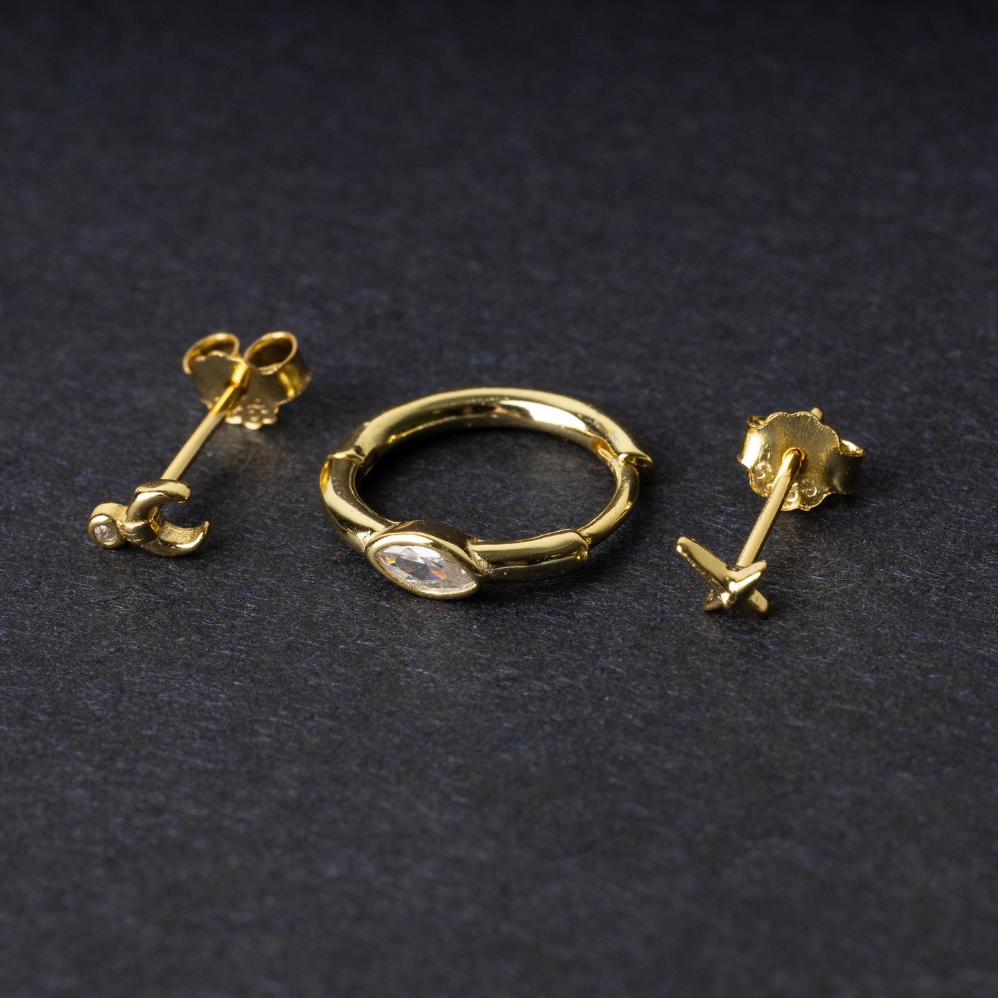 A gold-tone earring set displayed on a dark matte background, featuring a hoop with a marquise-cut crystal, a crescent moon stud with a crystal accent, and a minimalist cross stud.