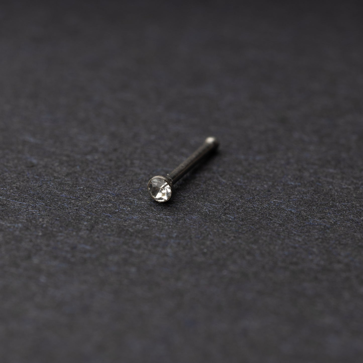 Stainless steel nose stud with a small crystal accent, displayed on a dark textured background.