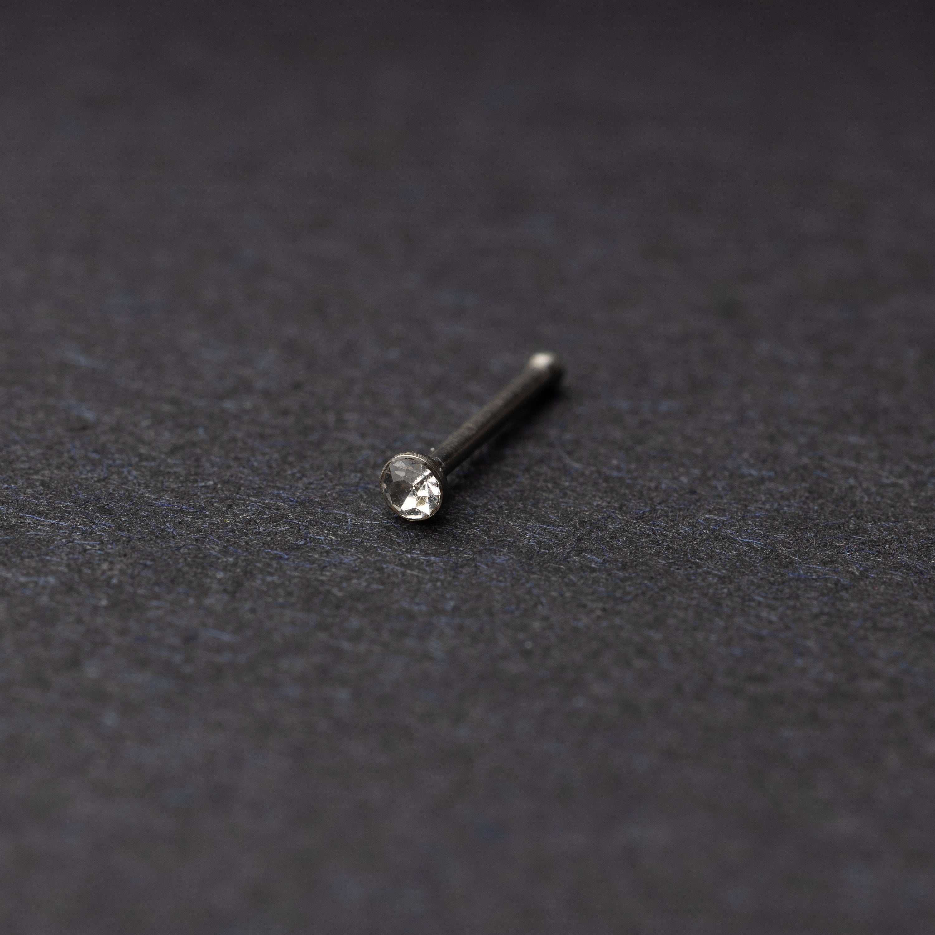 Stainless steel nose stud with a small crystal accent, displayed on a dark textured background.