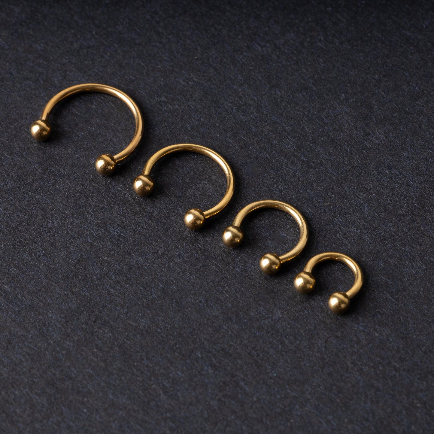 Set of four gold-colored titanium horseshoe rings with ball ends, displayed on a dark textured background.
