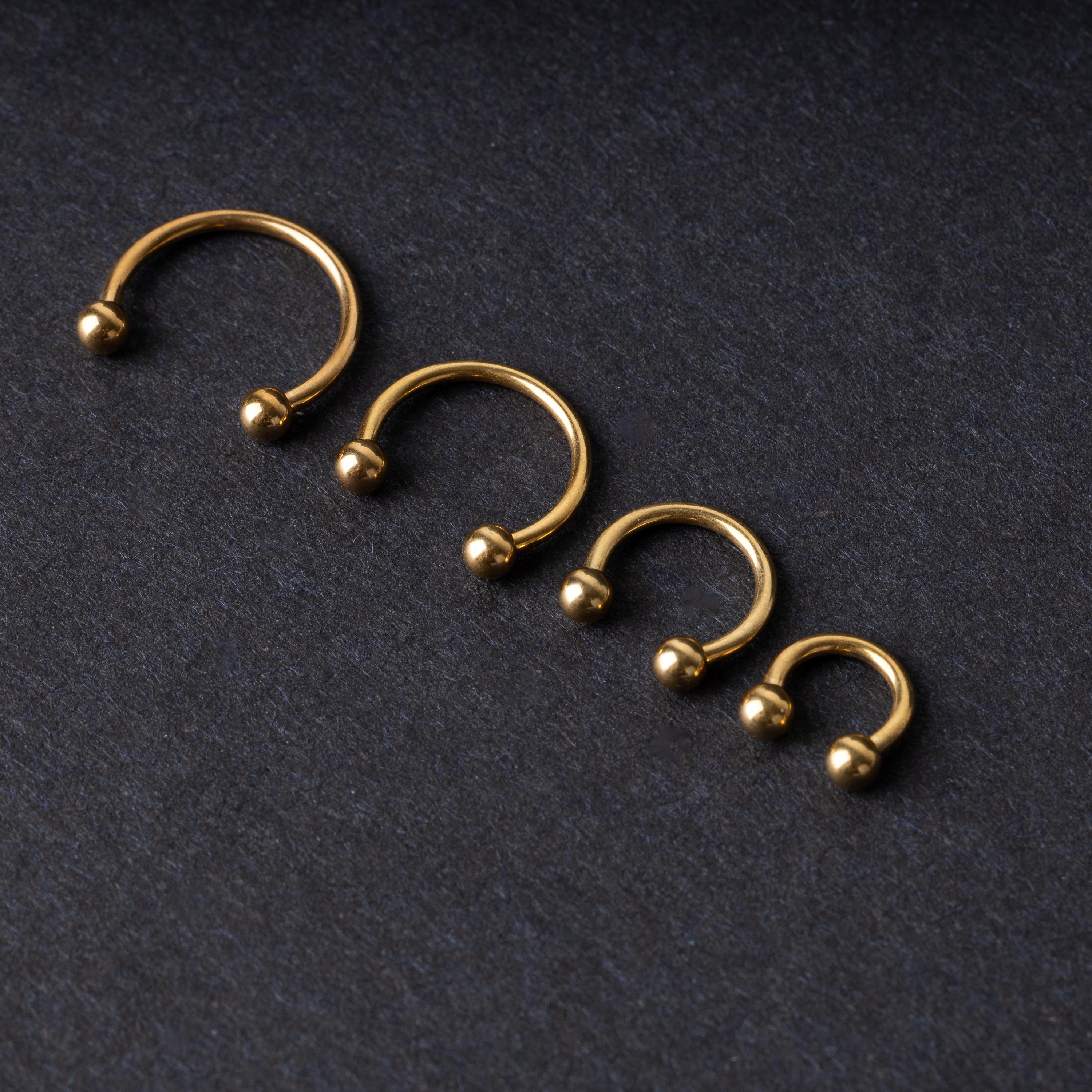 Set of four gold-colored titanium horseshoe rings with ball ends, displayed on a dark textured background.